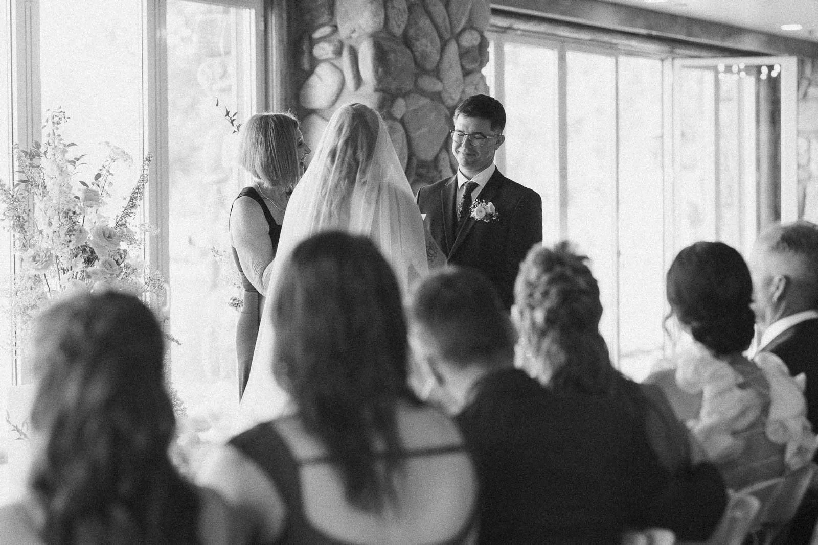  Facing each other at the altar, a bride and groom exchange vows with an officiant nearby, seated guests looking on in a light-filled, stone-accented venue. 