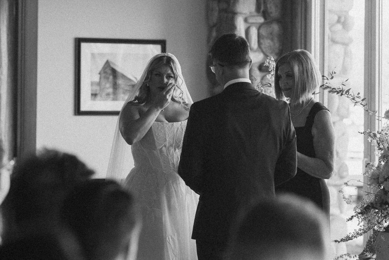  An emotional bride dabs away tears beside the groom and officiant at an indoor ceremony, sunlight pouring in through tall windows. 