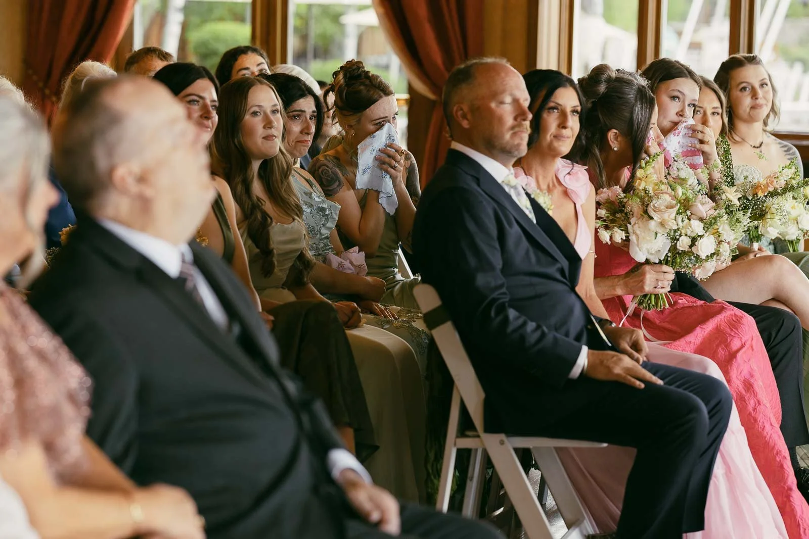  Wedding guests sit indoors, dressed formally and attentively watching an event. Some women hold bouquets and tissues, suggesting emotional moments. Large windows and curtains are visible in the background, bringing in natural light. 