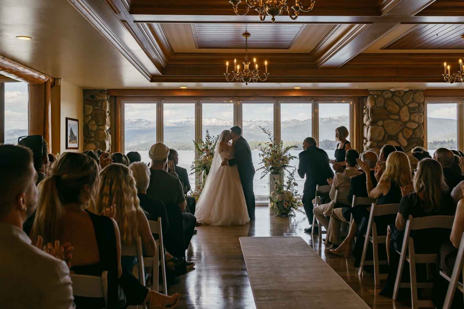  A bride and groom share a kiss at the altar in a rustic indoor wedding ceremony, with guests seated and mountains visible through large windows behind them. 