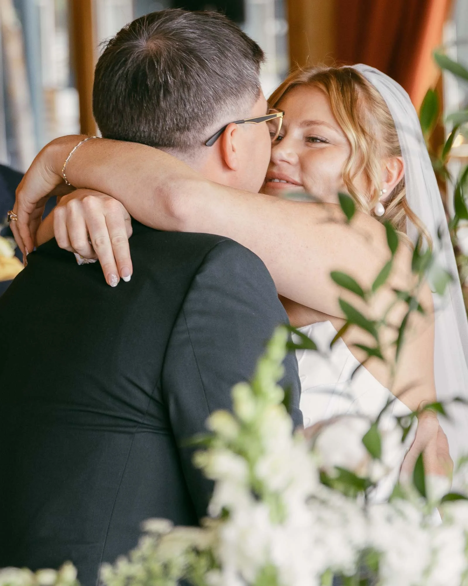  A bride and groom embrace closely, smiling at each other, surrounded by greenery and white flowers at what appears to be their wedding reception. 