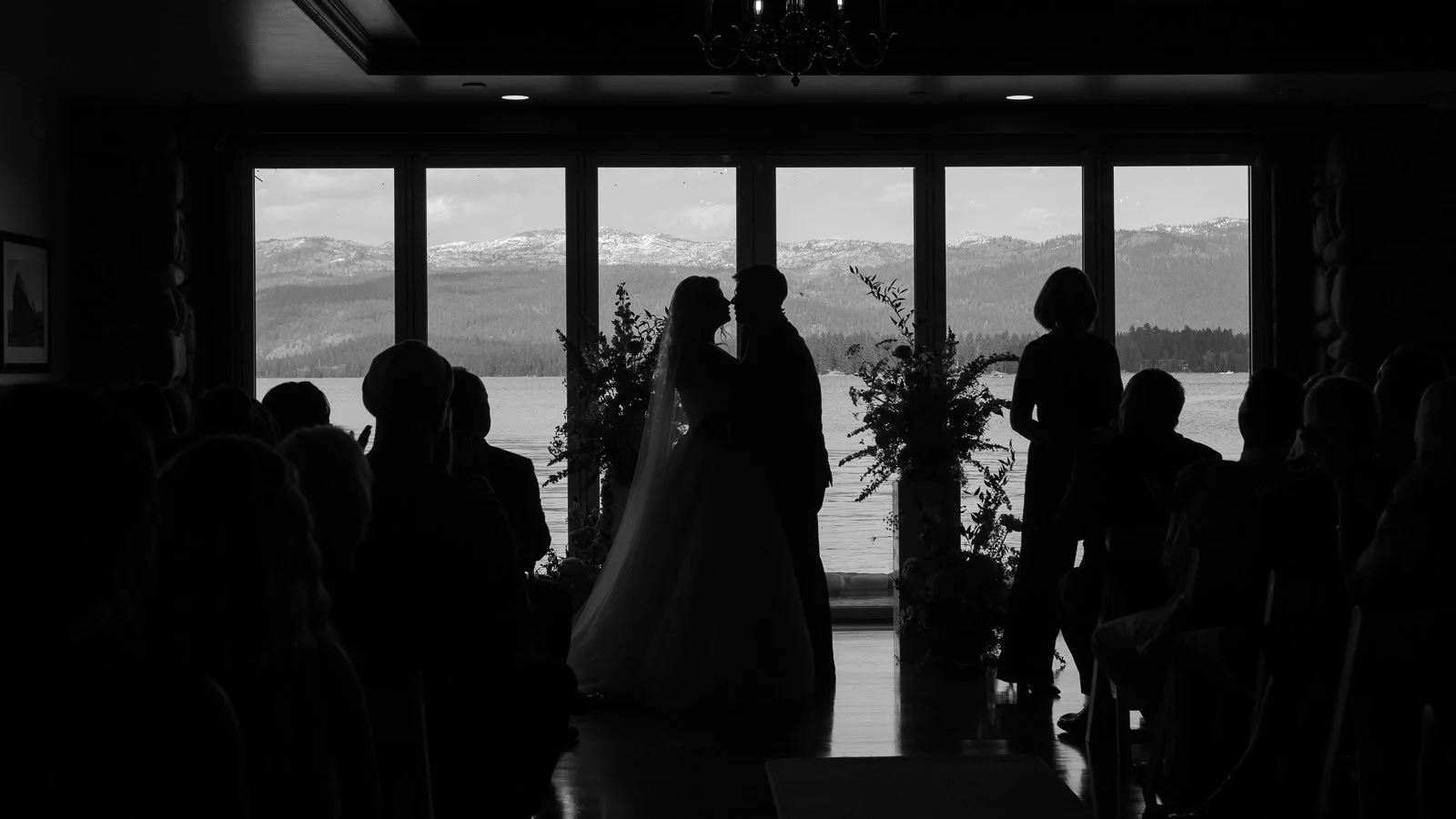  Silhouetted couple kisses at their wedding in front of large windows overlooking a lake and mountains, surrounded by guests. 