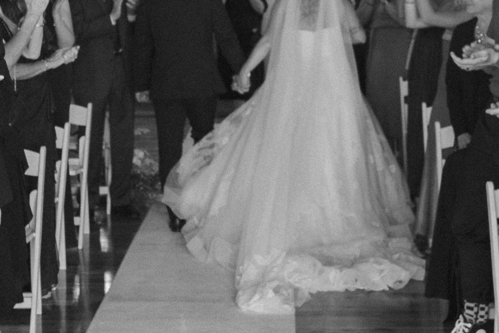  A bride and groom walk hand in hand down an aisle lined with seated guests, the bride wearing a long, flowing dress and veil. 