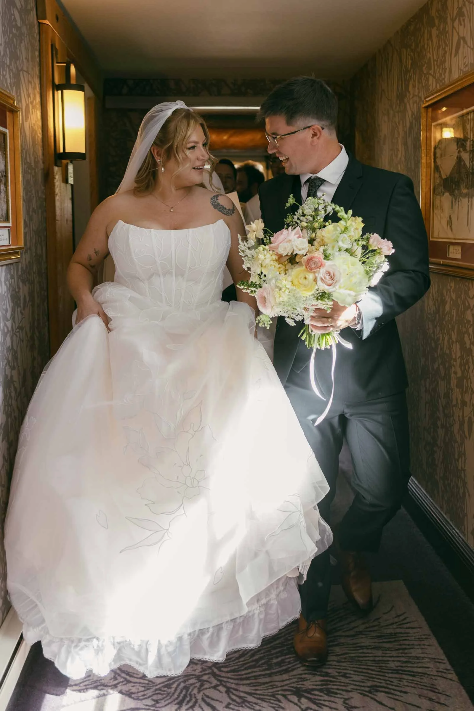  A bride in a white gown holds her dress while walking with a man in a suit, who carries a bouquet of flowers. They smile at each other in a warmly lit hallway. 