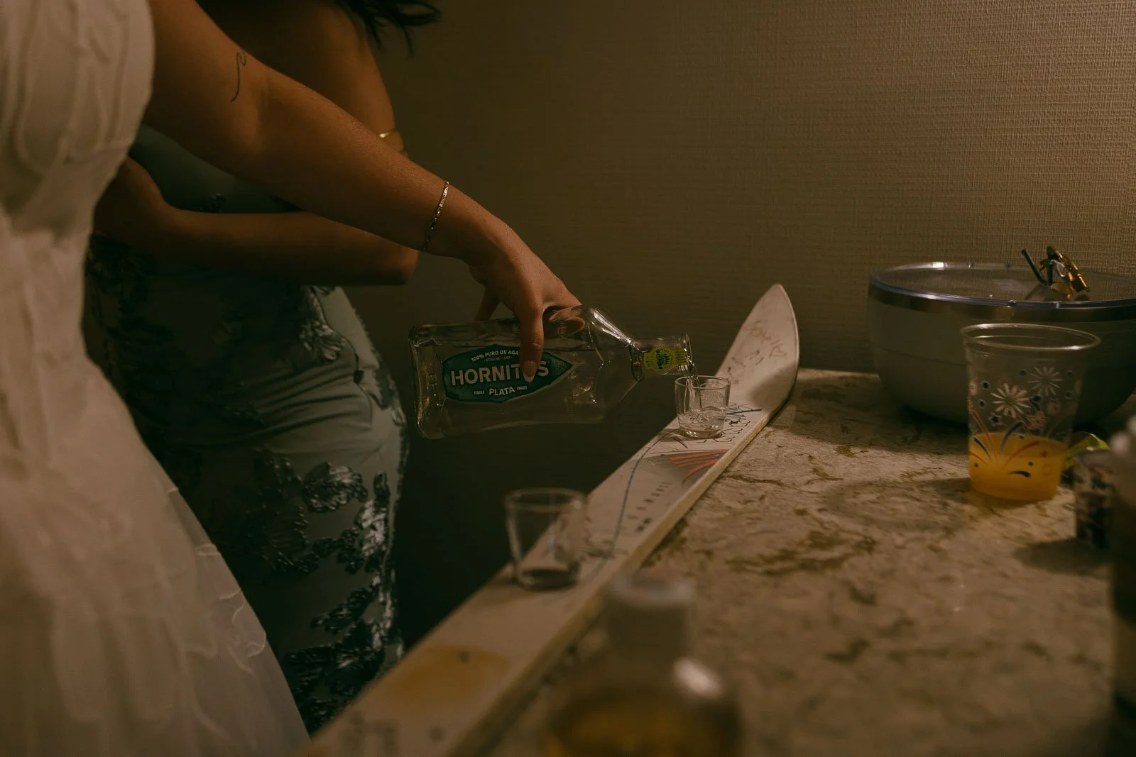  The bride pours Hornitos tequila into shot glasses lined up on a ski, with drinks and a bowl on a counter nearby. 