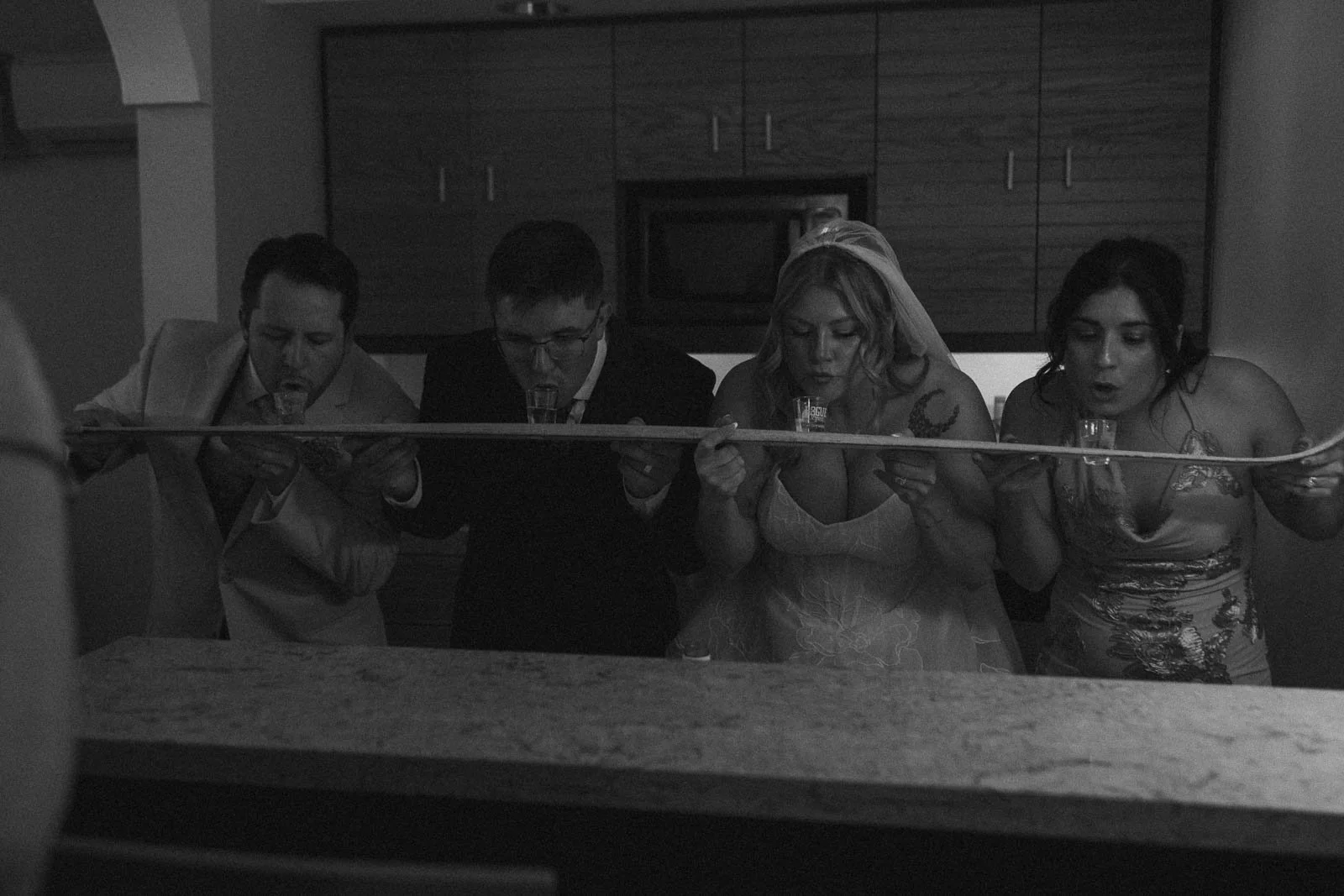  Four people, including a bride in a wedding dress, lean over a counter to take a group shot from a wooden board holding shot glasses. The image is in black and white. 