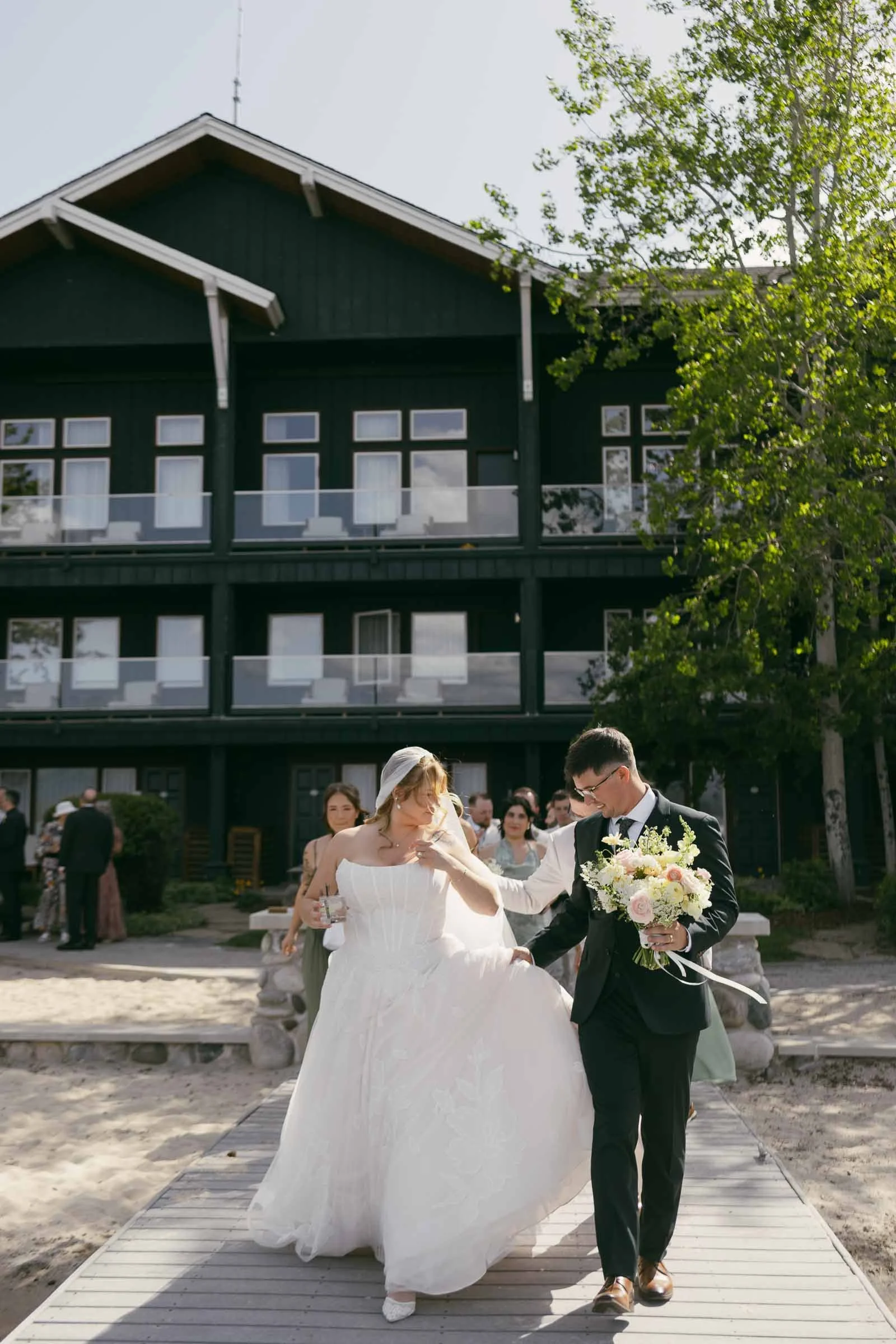  A bride and groom walk outside on a sunny day, the bride holding her dress and the groom carrying a bouquet, with guests and a dark building in the background. 