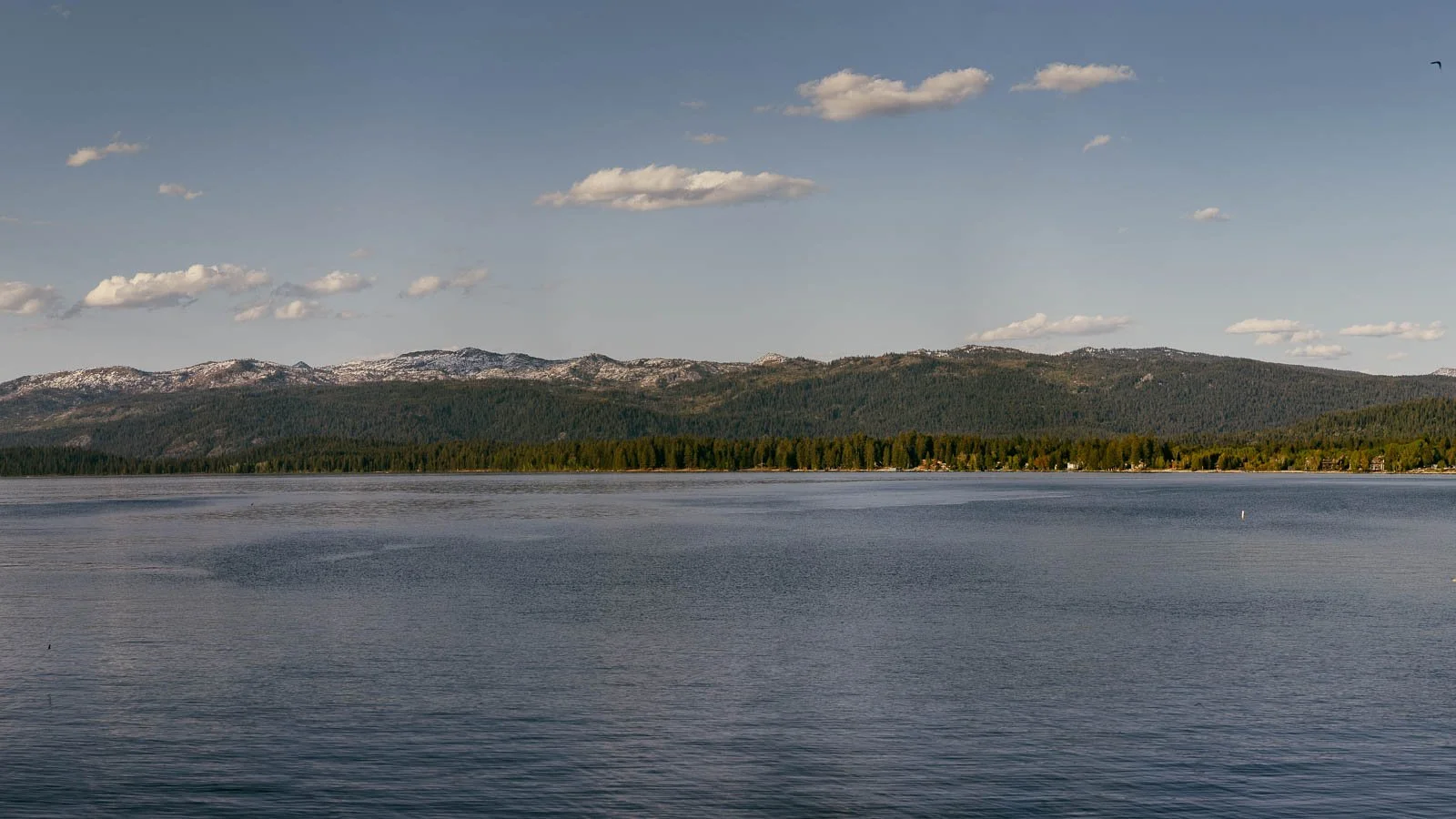  A calm lake stretches across the foreground, reflecting the sky. In the background, forested mountains rise under a partly cloudy blue sky. 