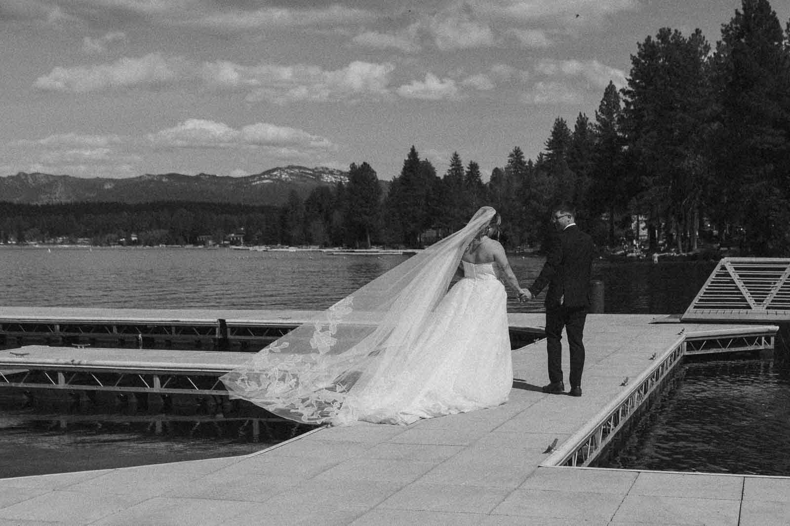  A bride and groom walk hand-in-hand on a dock by a lake, surrounded by trees and mountains, under a partly cloudy sky. 