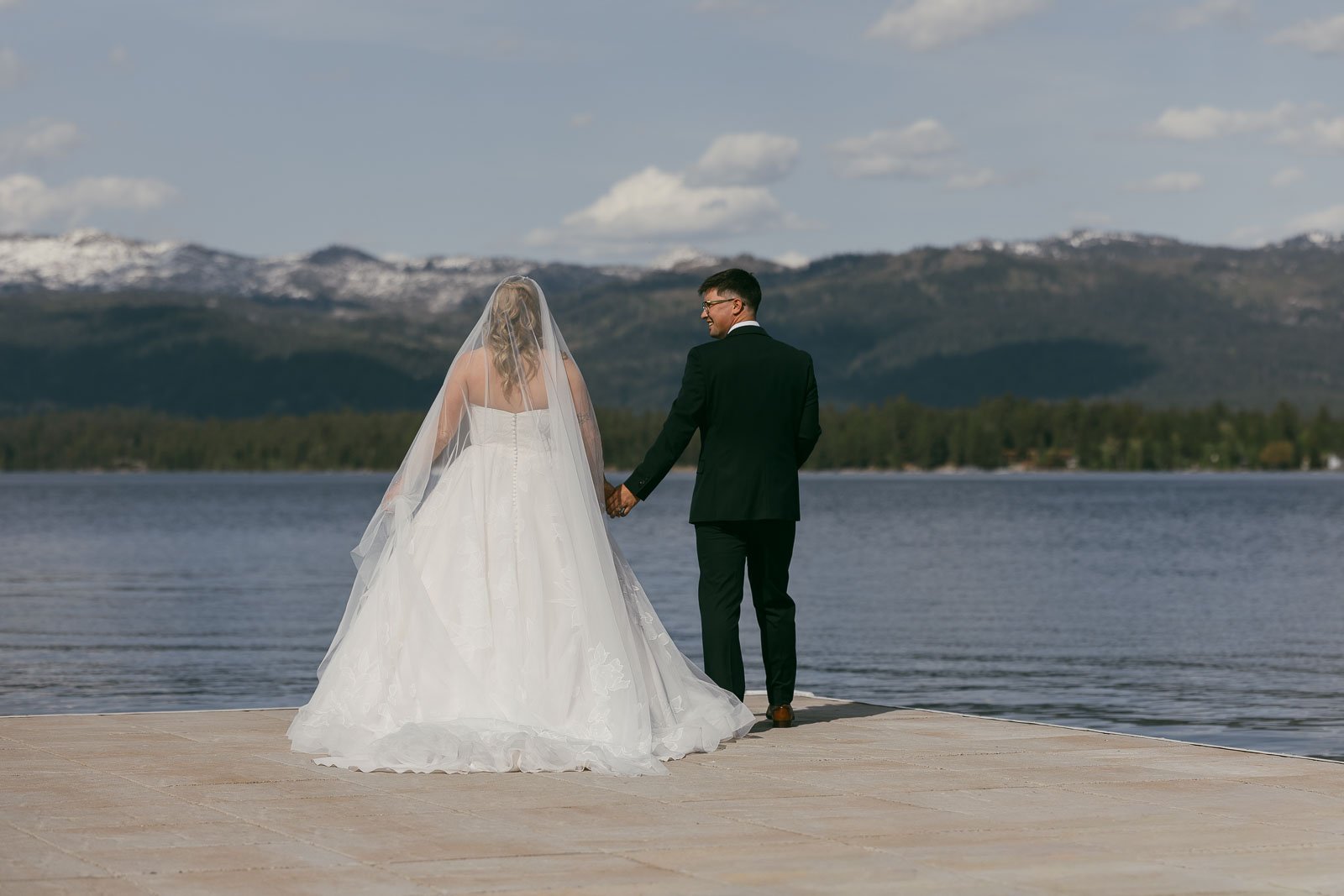 A bride and groom stand hand in hand on a dock, facing a calm lake with mountains and trees in the background under a blue sky. 