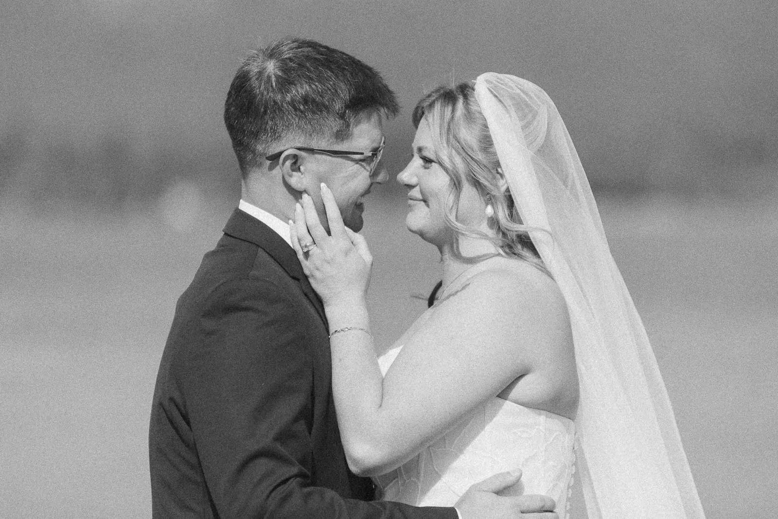  A bride and groom face each other, smiling and touching foreheads. The bride gently holds the groom's face, and both wear wedding attire outdoors on a sunny day. 