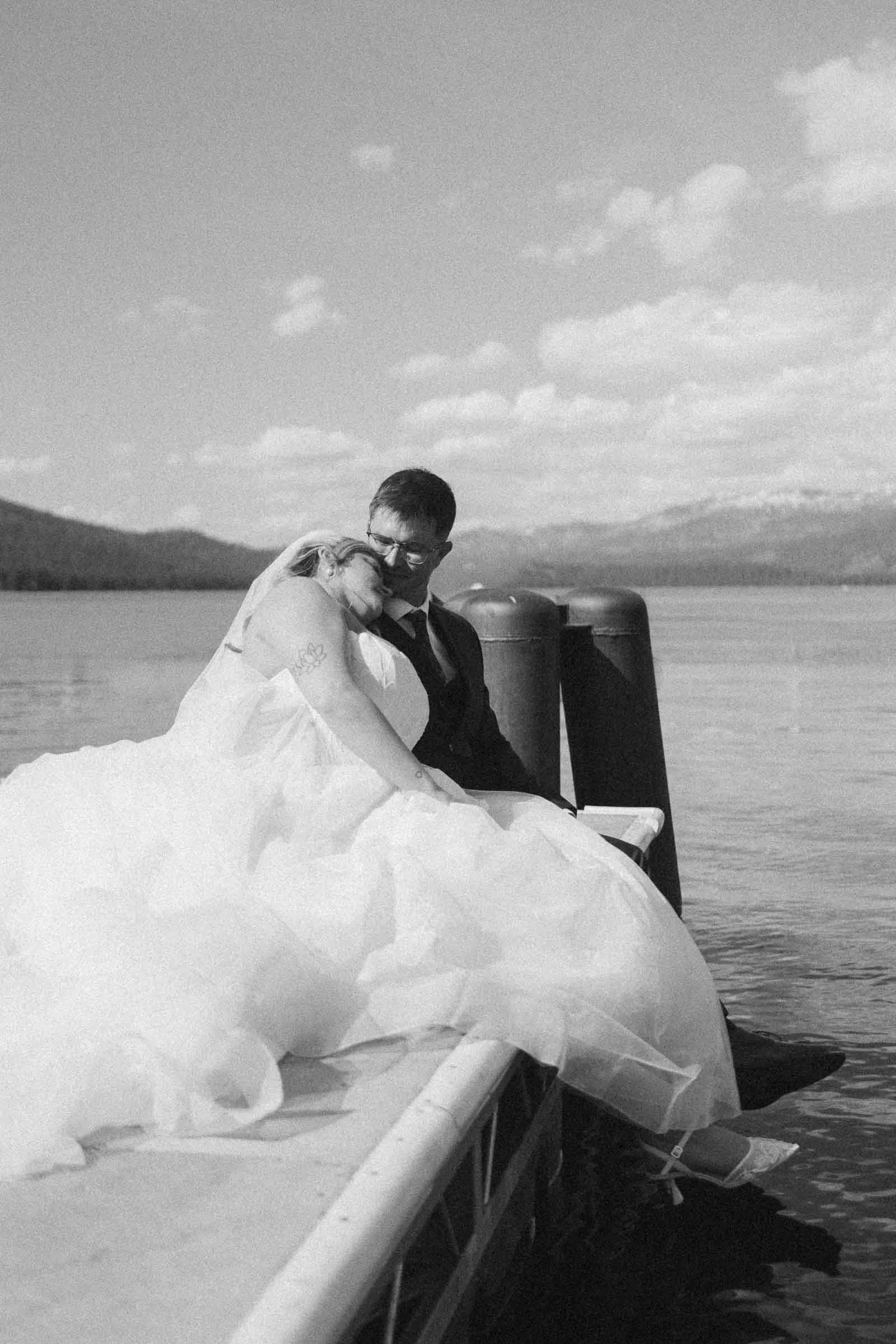  A bride and groom sit at the edge of a dock by a lake, with mountains in the background. The bride rests her head on the groom’s shoulder. The image is in black and white. 