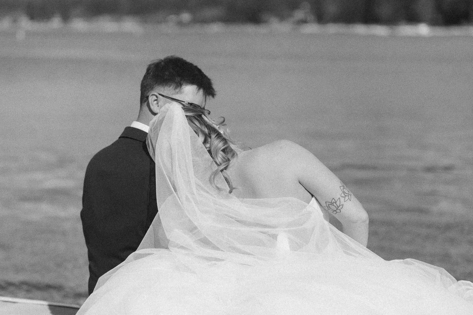  A bride in a strapless dress and veil sits beside a groom in a suit, both facing away, by a body of water. 