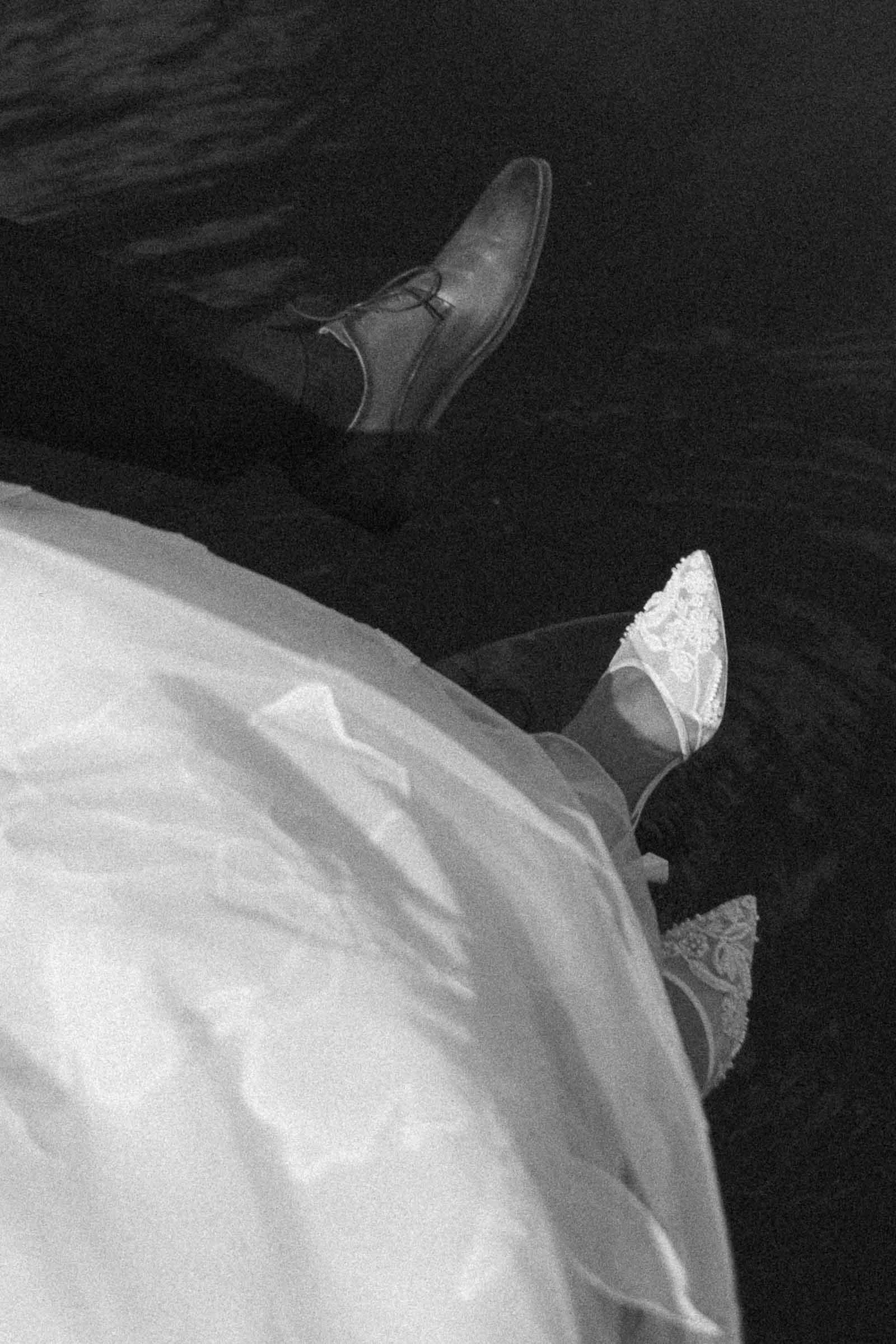  A black-and-white photo showing a bride in a wedding dress and lace shoes sitting beside someone in dress shoes, both with their feet over the water. 