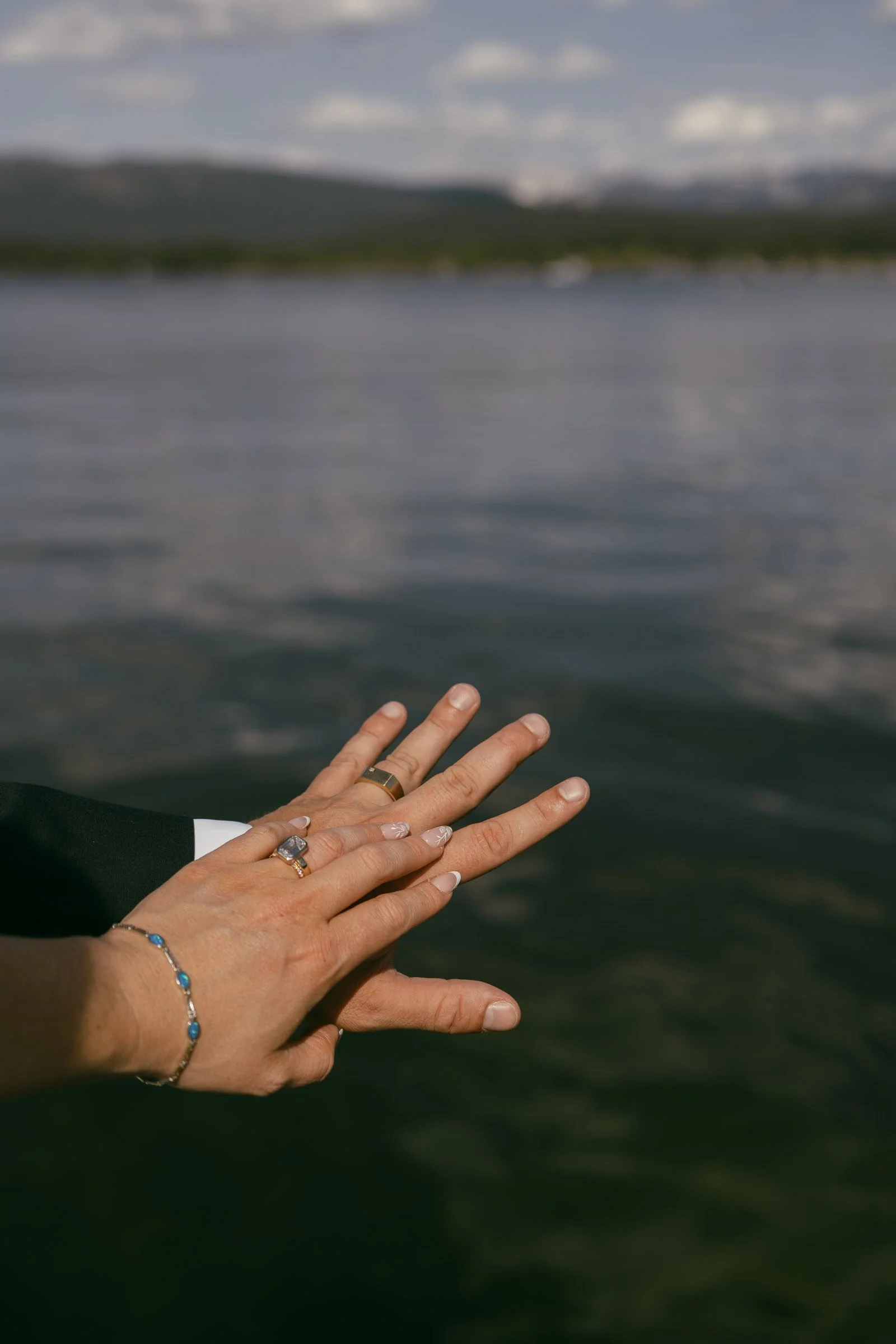  Two hands with wedding rings rest together, fingers extended, against a blurred backdrop of calm water and distant shoreline, suggesting a romantic or celebratory moment. 