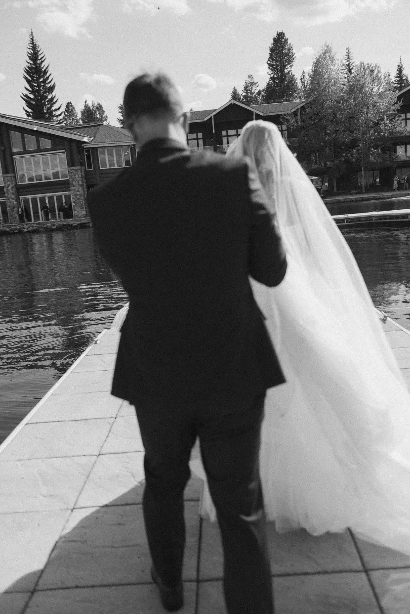  A bride and groom walk together on a dock by the water, with houses and trees in the background. The image is in black and white and taken from behind them. 