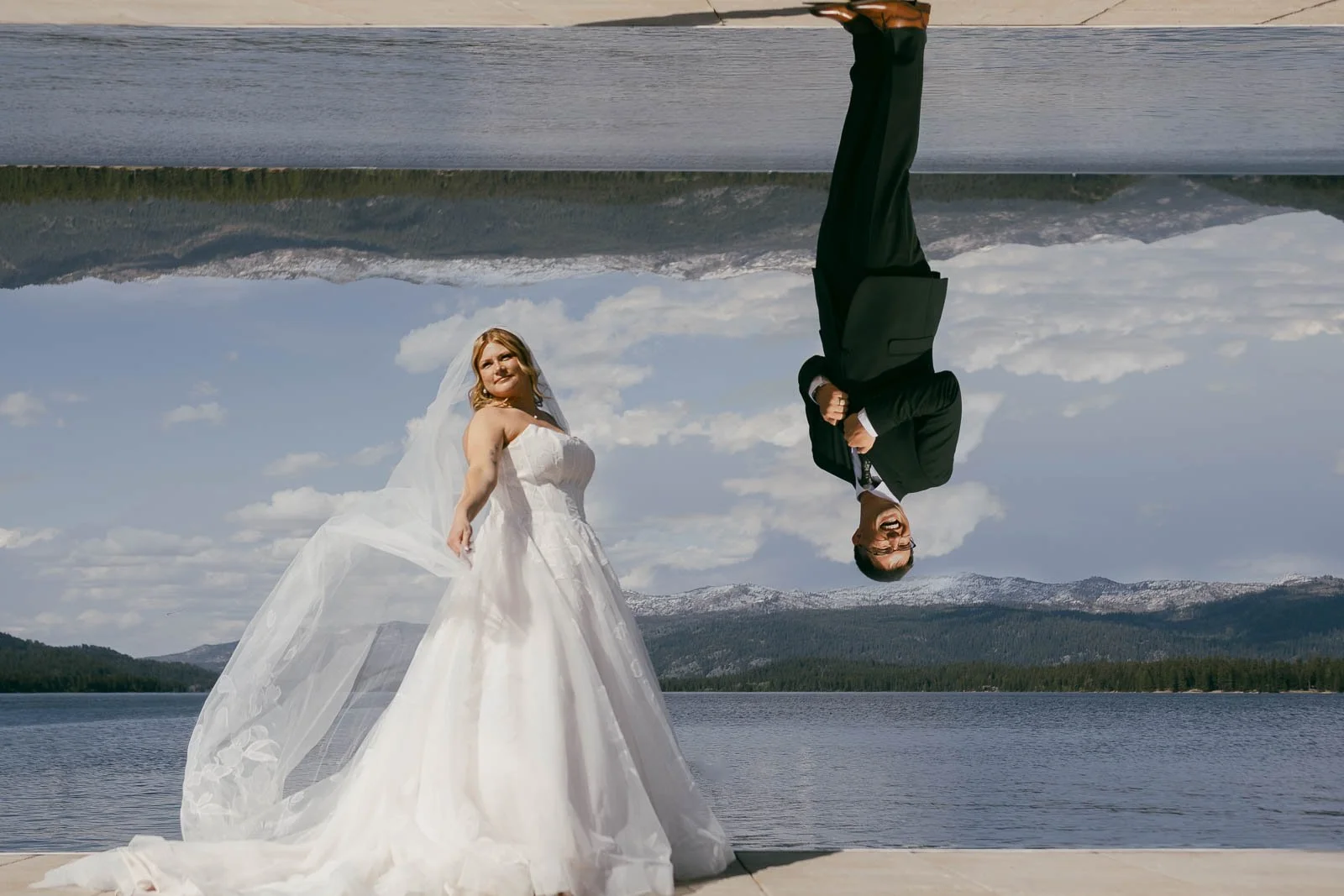  A bride in a white gown poses by a lake with mountains behind her. Above her is a flipped reflection of a groom in a black suit, creating an upside-down effect. The scene is bright with a clear sky. 