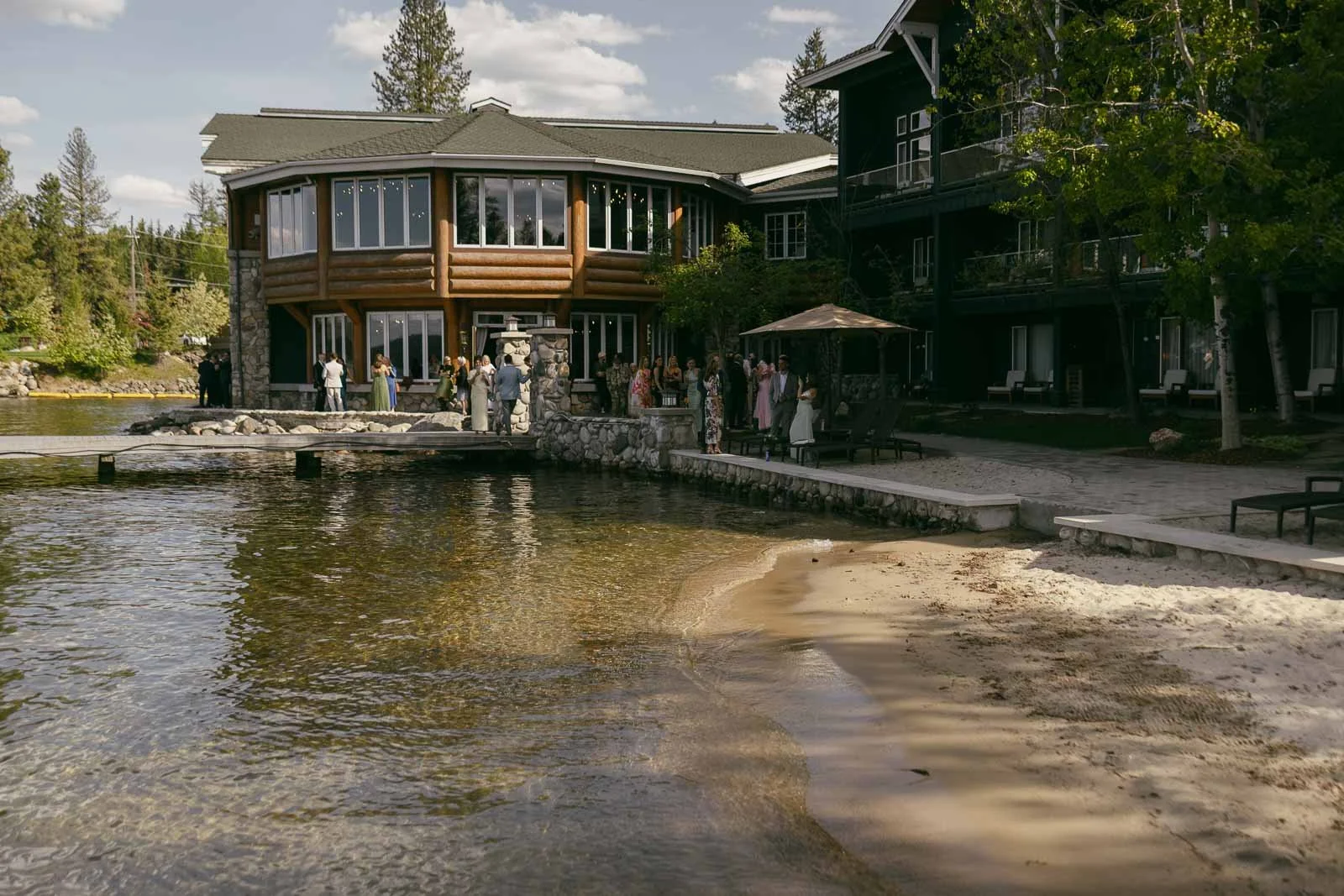  A group of people stands near the edge of a rustic lakeside lodge with large windows. There is a sandy beach, clear water, trees, and a partly cloudy sky. 
