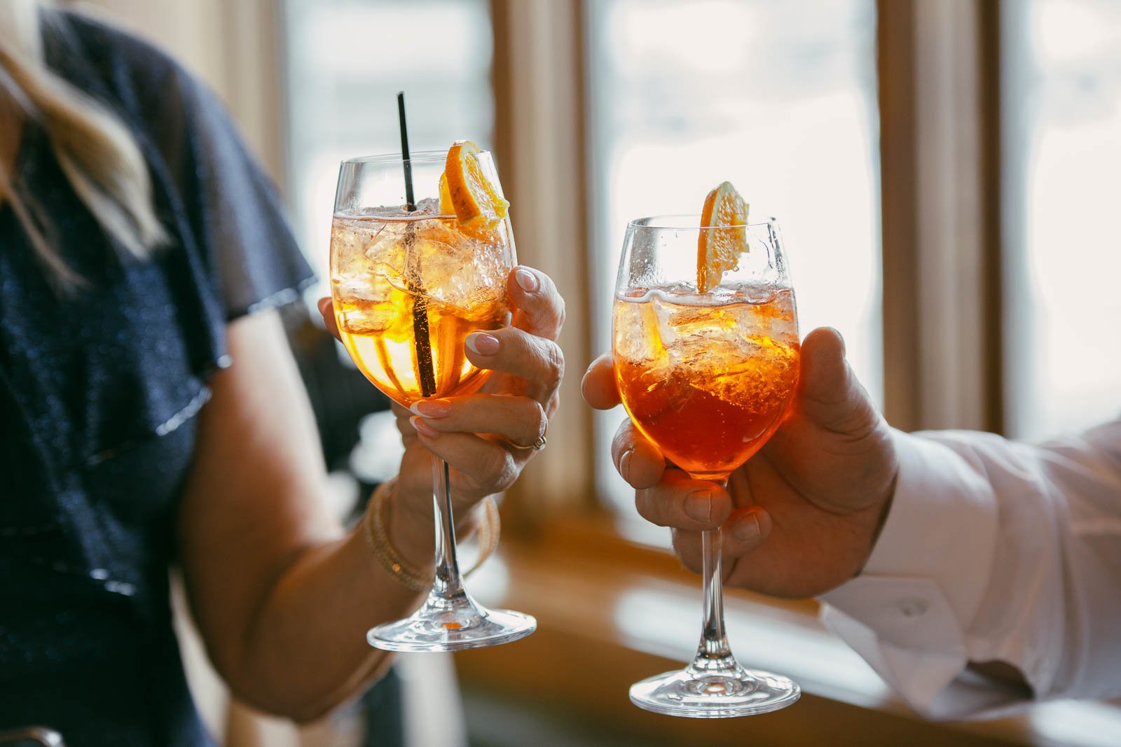  Two people clink glasses filled with orange cocktails, ice, and orange slices. The background shows blurred windows and sunlight. 
