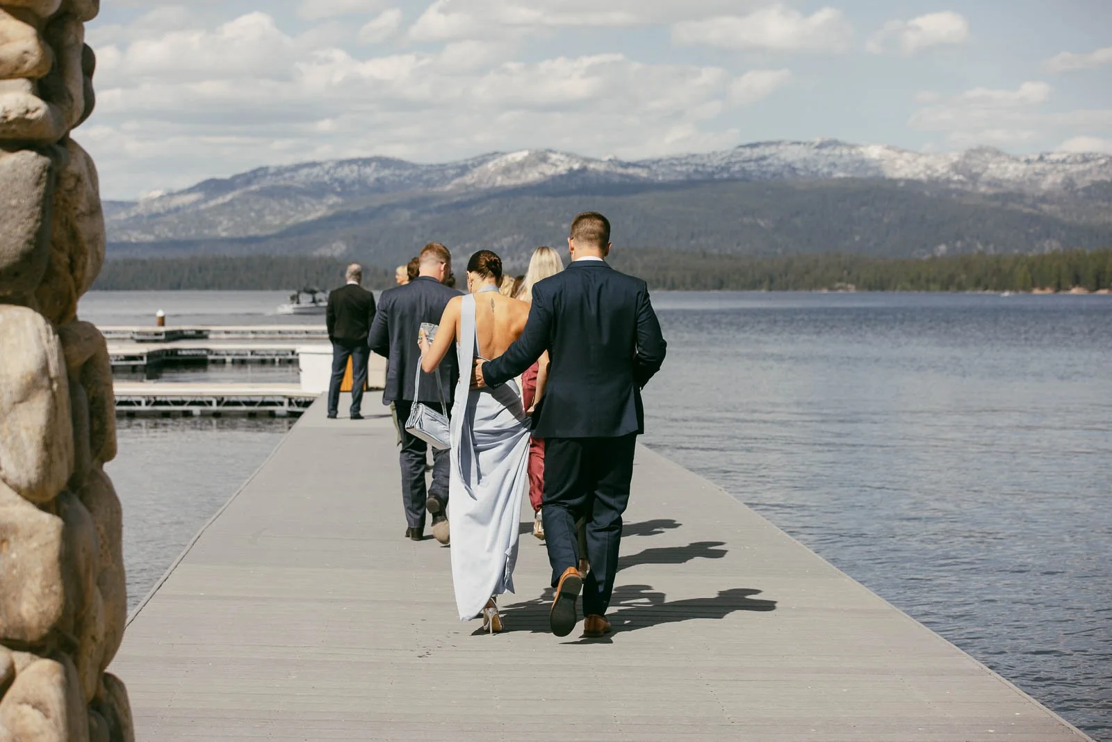  A group of people dressed formally walk down a dock by a lake, with mountains and trees in the background under a partly cloudy sky. 