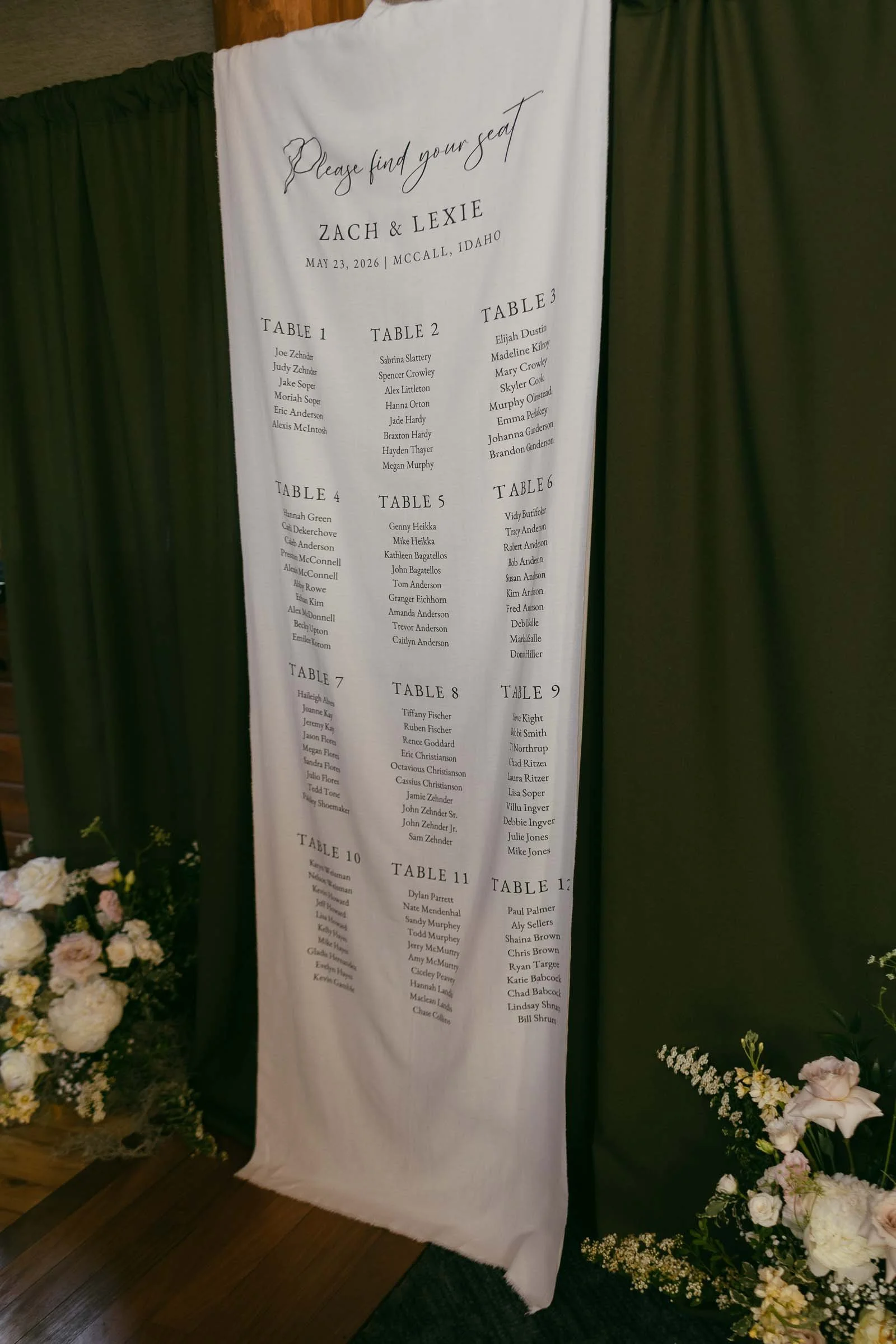  A white wedding seating chart for Zach and Lexie, dated May 23, 2026, in McCall, Idaho, hangs between green curtains, surrounded by white and pink floral arrangements. 