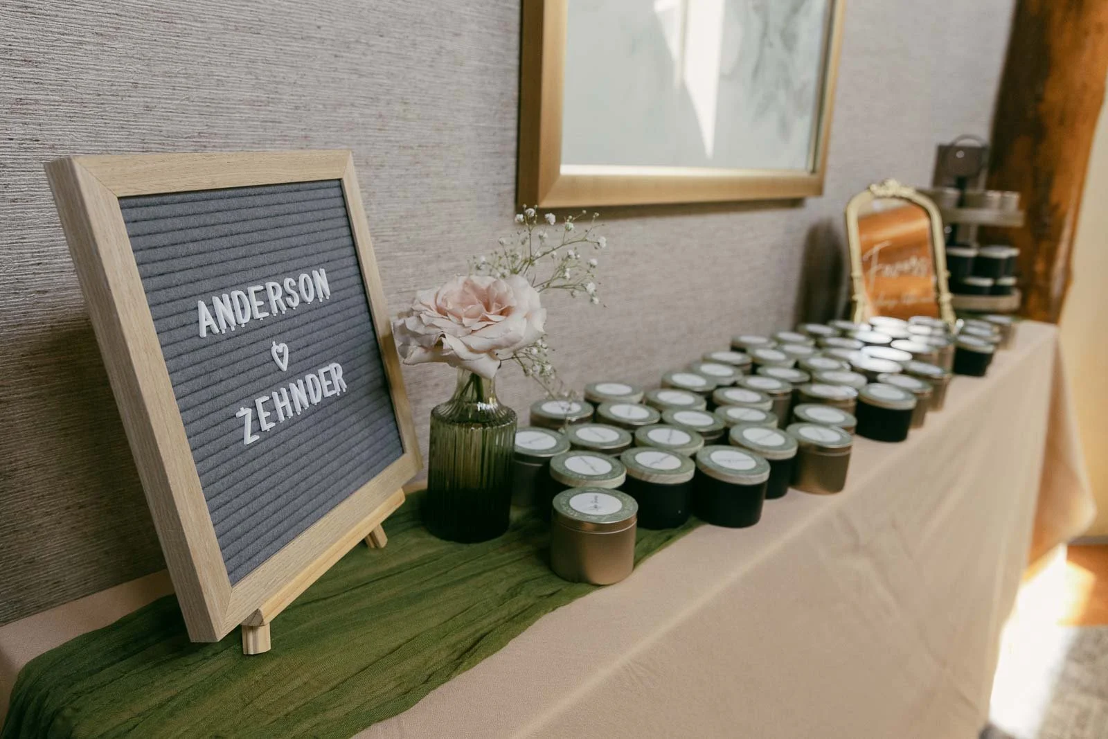  A table holds many small candles, a framed sign reading "Anderson ♥ Zehnder," a pink rose in a vase, and a mirror with cursive writing, all arranged for a wedding or event. 
