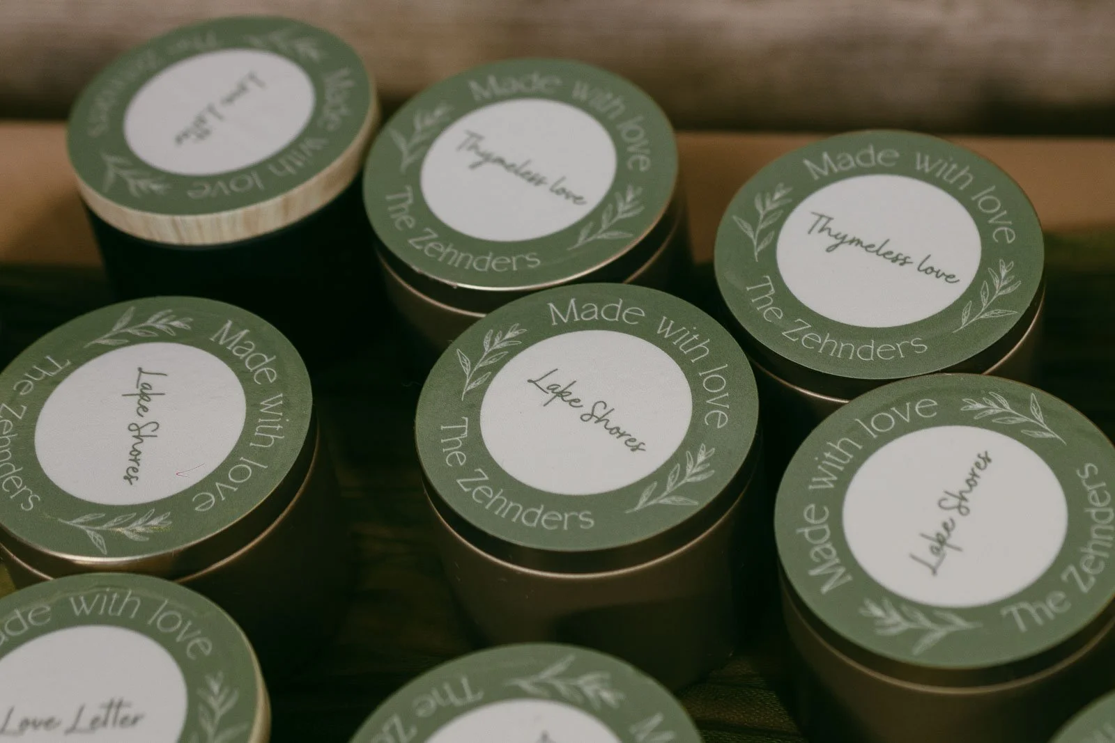  Round containers with green lids labeled "Made with love The Zehnders," some also say "Thymeleaf love" and "Lake Shower," arranged closely together on a surface. 