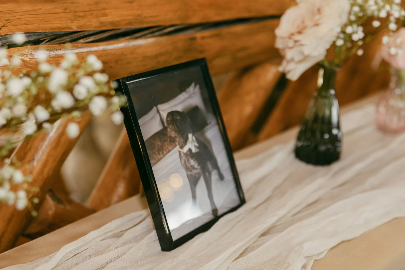  A framed photo of a dog is displayed on a light cloth-covered table, surrounded by vases with flowers and baby's breath, against a wooden background. 