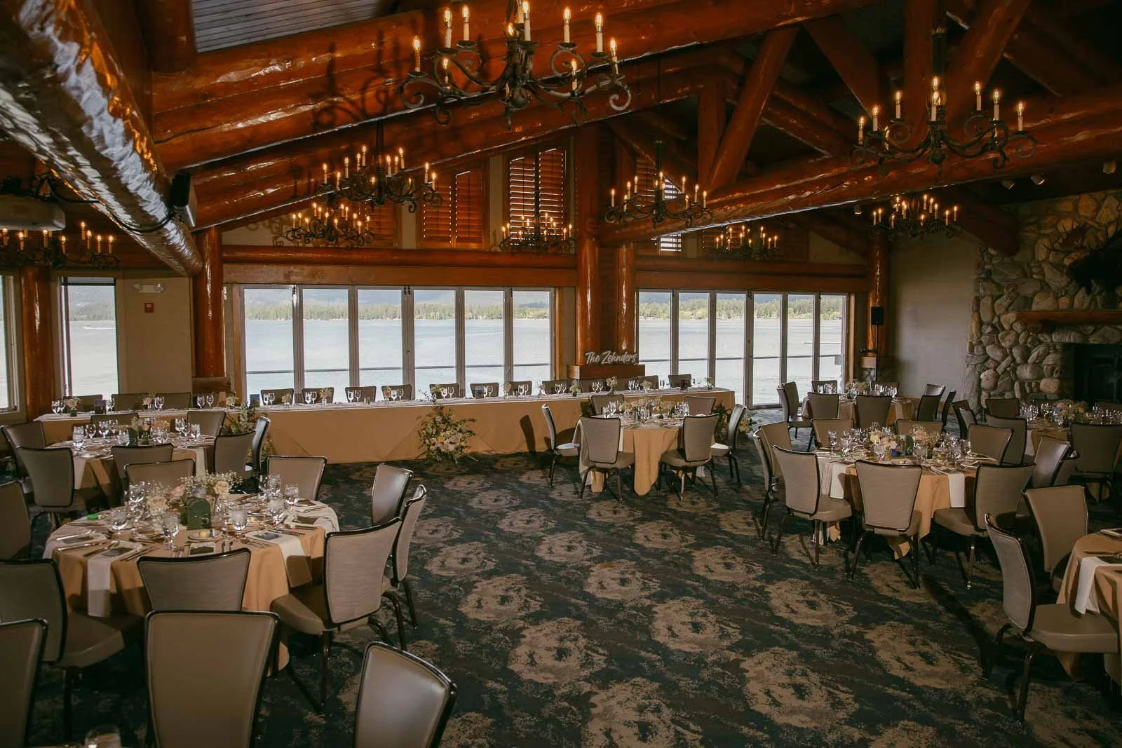  Rustic banquet hall with log beams, chandeliers, round tables set for an event, large windows showing a lake and trees outside, and a stone fireplace on the right. 