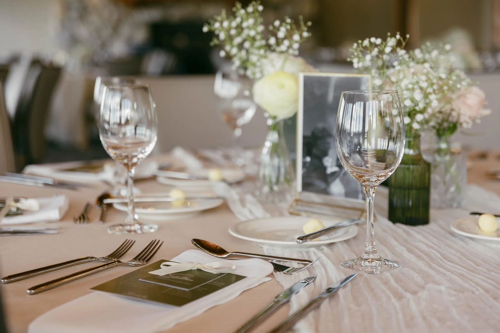  Elegant table setting with wine glasses, plates, silverware, white napkins, menu cards, and small flower arrangements in vases on a beige tablecloth. The scene is prepared for a formal event or celebration. 