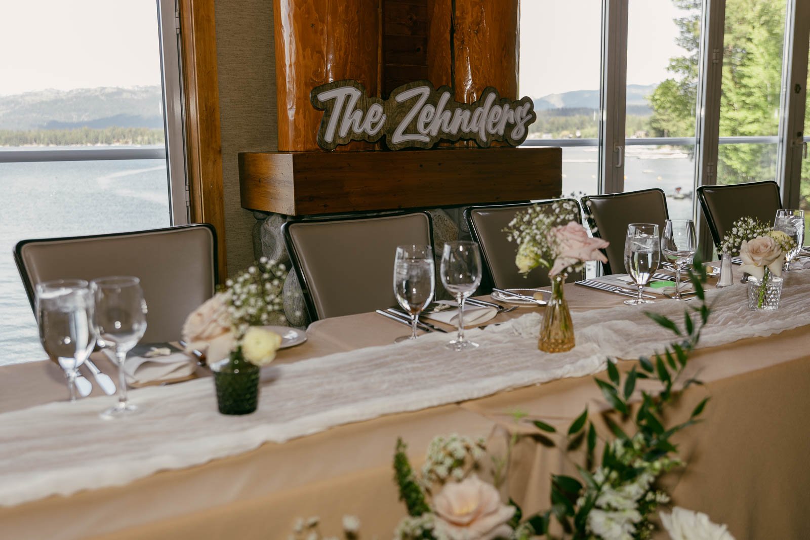  Elegant wedding table set with flowers, glassware, and napkins, overlooking a lake through large windows. A sign reading "The Zehnders" is displayed on a mantel behind the table. 