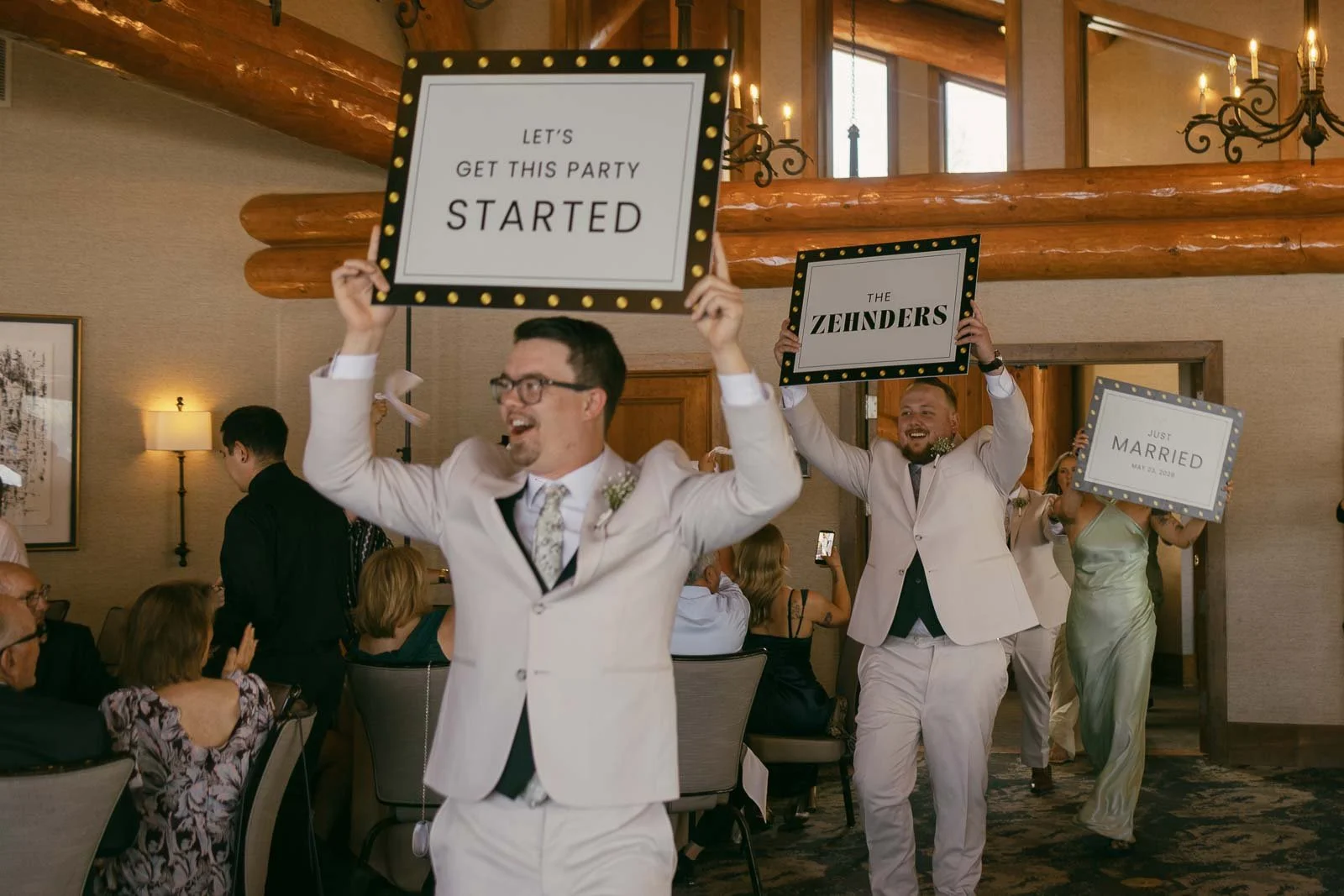  Two men in light suits hold up signs reading "LET'S GET THIS PARTY STARTED" and "THE ZEHNDERS." People are seated around them, smiling, in a warmly lit, wood-beamed event space. 