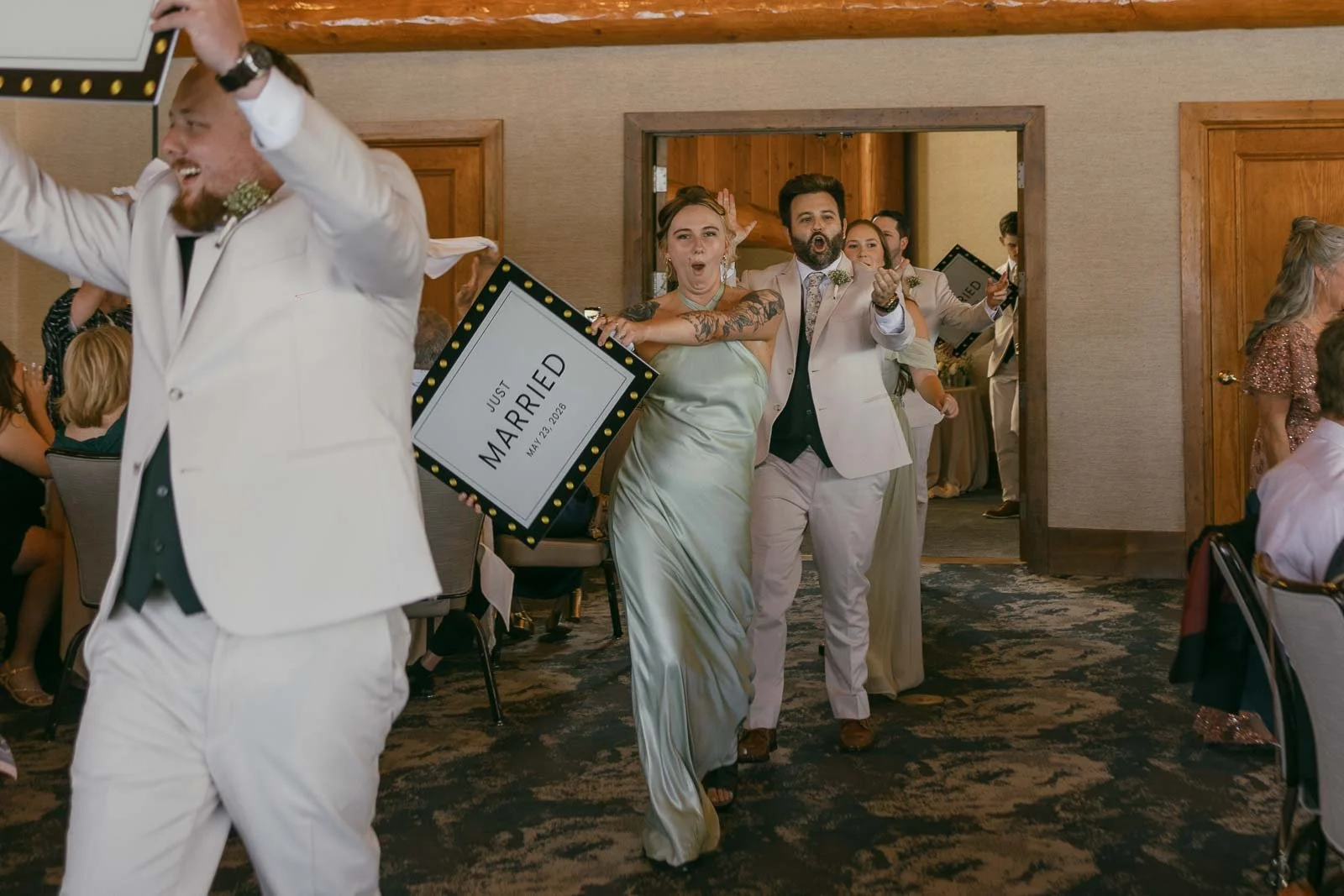  A group of people in formal attire excitedly enter a room holding "Just Married" signs, celebrating a wedding. 