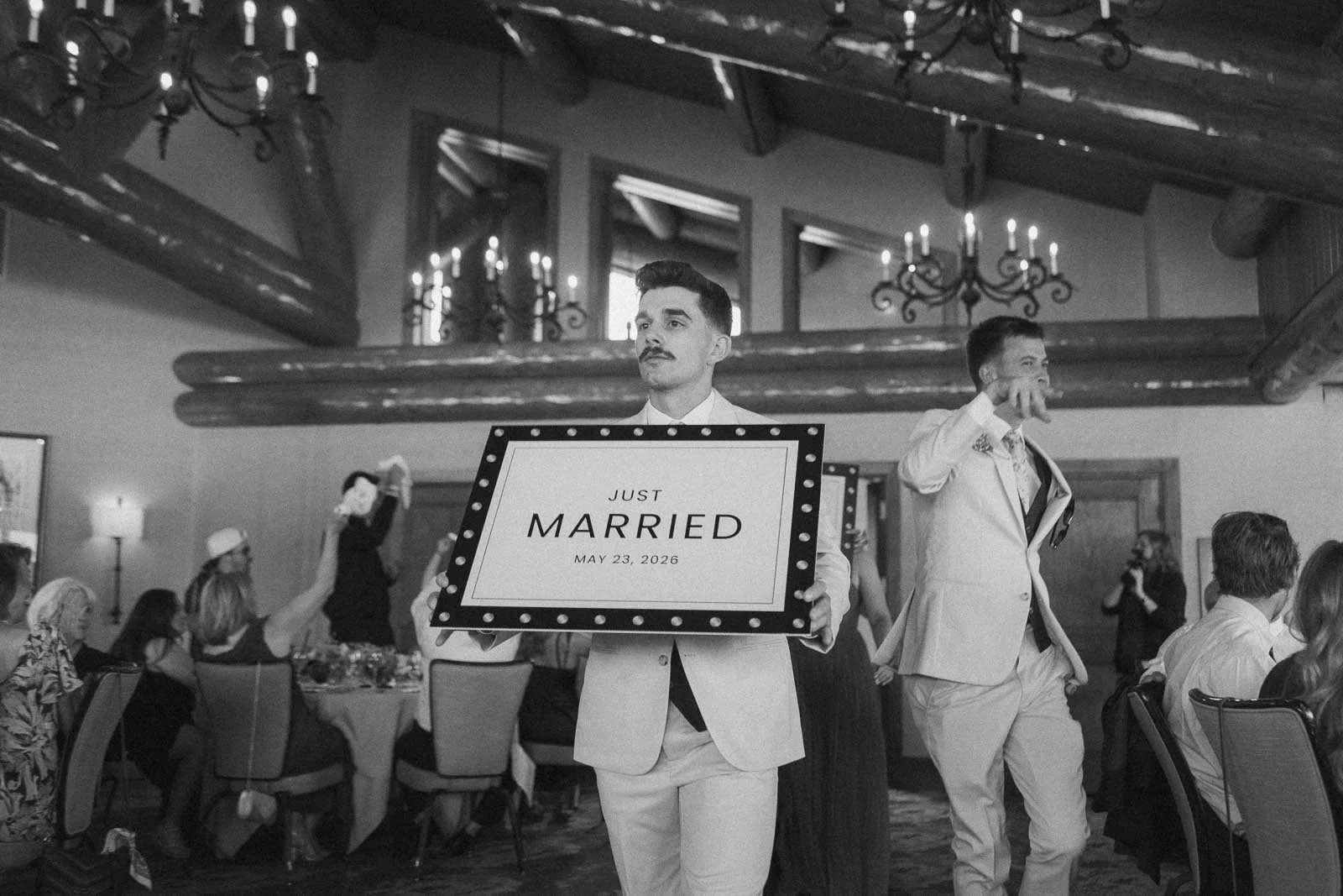  A man in a suit holds a sign reading "Just Married May 23, 2026" at a formal indoor event, while guests seated at tables watch. Chandeliers hang from the ceiling. 