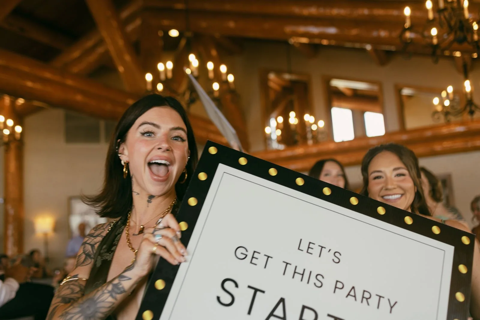  Two smiling women hold a large sign reading “LET’S GET THIS PARTY STARTED” in a warmly lit room with chandeliers and wooden beams, creating a festive, joyful atmosphere. 