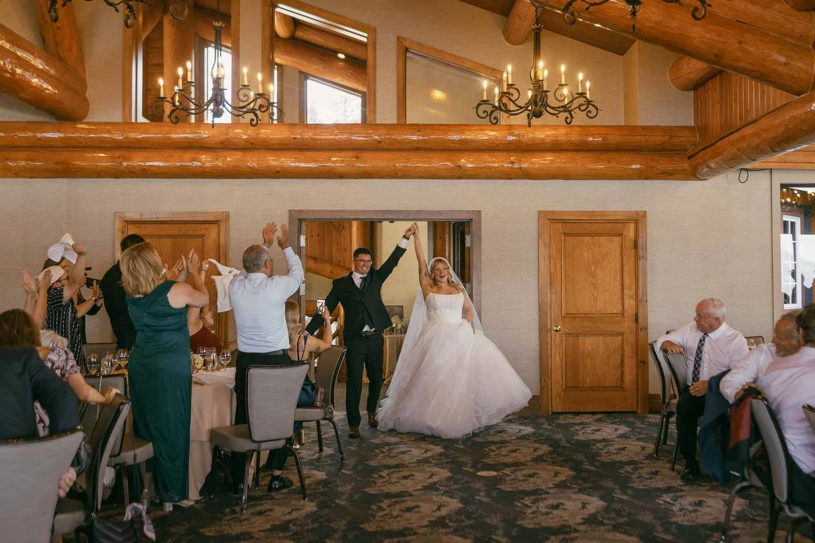  A bride and groom enter a rustic reception hall, holding hands and smiling as guests cheer and clap around them. The room features wooden beams, chandeliers, and round tables. 