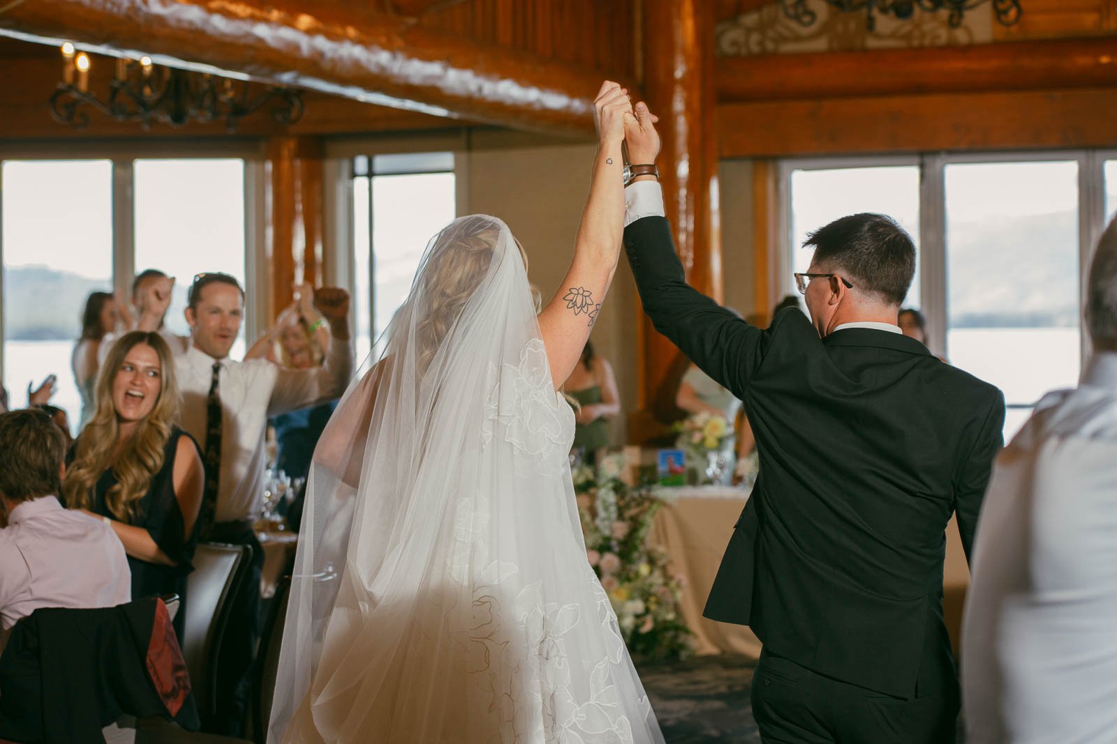  A bride and groom, holding hands, raise their arms as they walk into a reception with cheering guests around them. The bride wears a veil, and the groom is in a suit. The atmosphere is joyful and celebratory. 