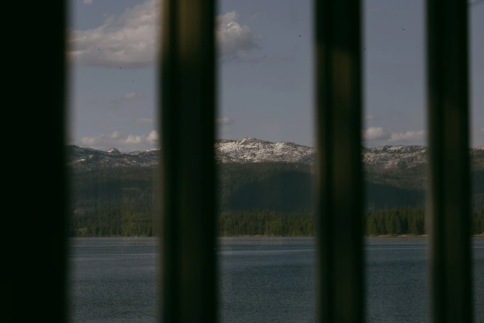  Snow-capped mountains and a lake are visible through the gaps between vertical dark bars, with a forested shoreline in the background under a partly cloudy sky. 