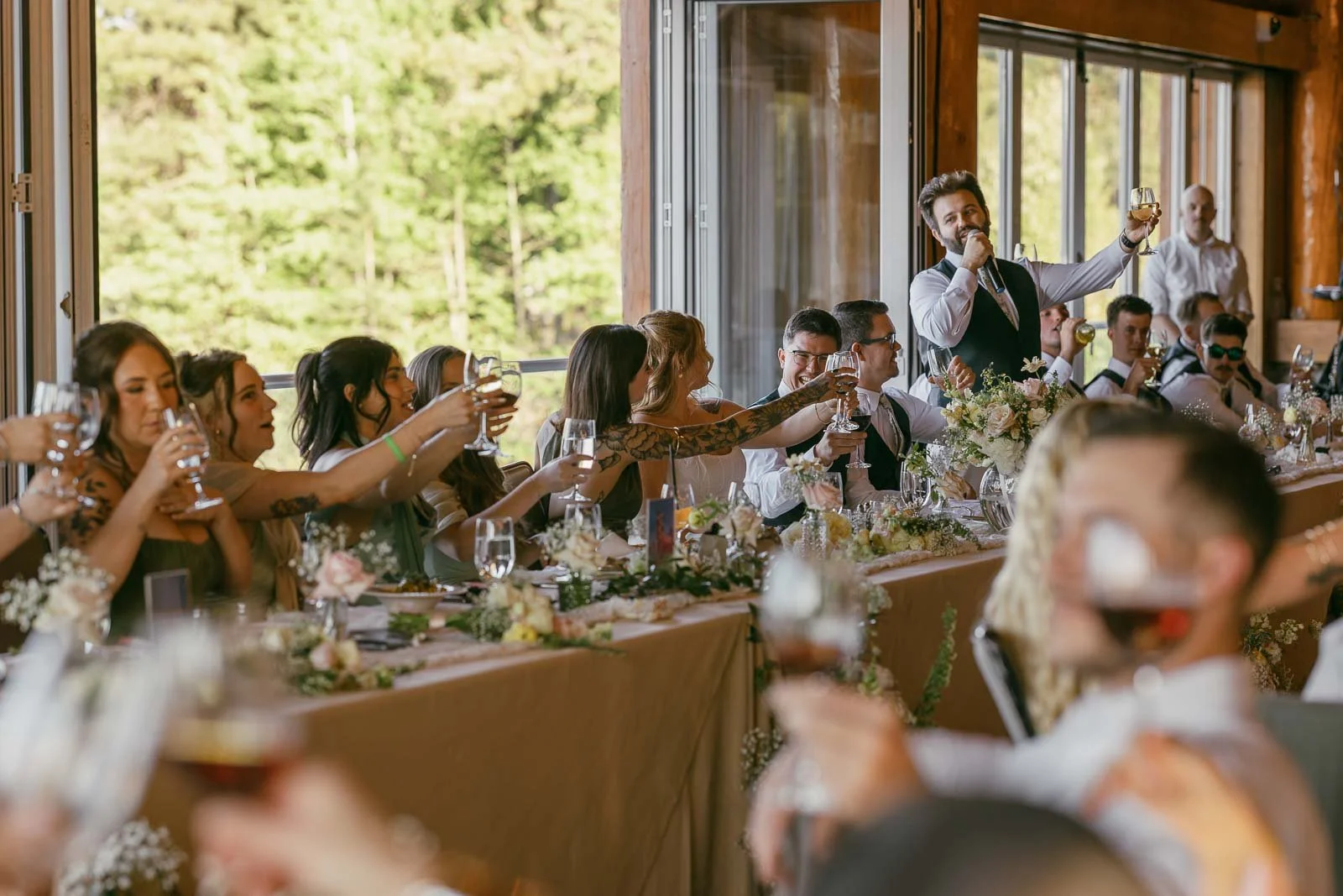  A group of people seated at long tables raise their glasses in a toast during a celebration, with a man standing and speaking, surrounded by floral decorations and natural light. 