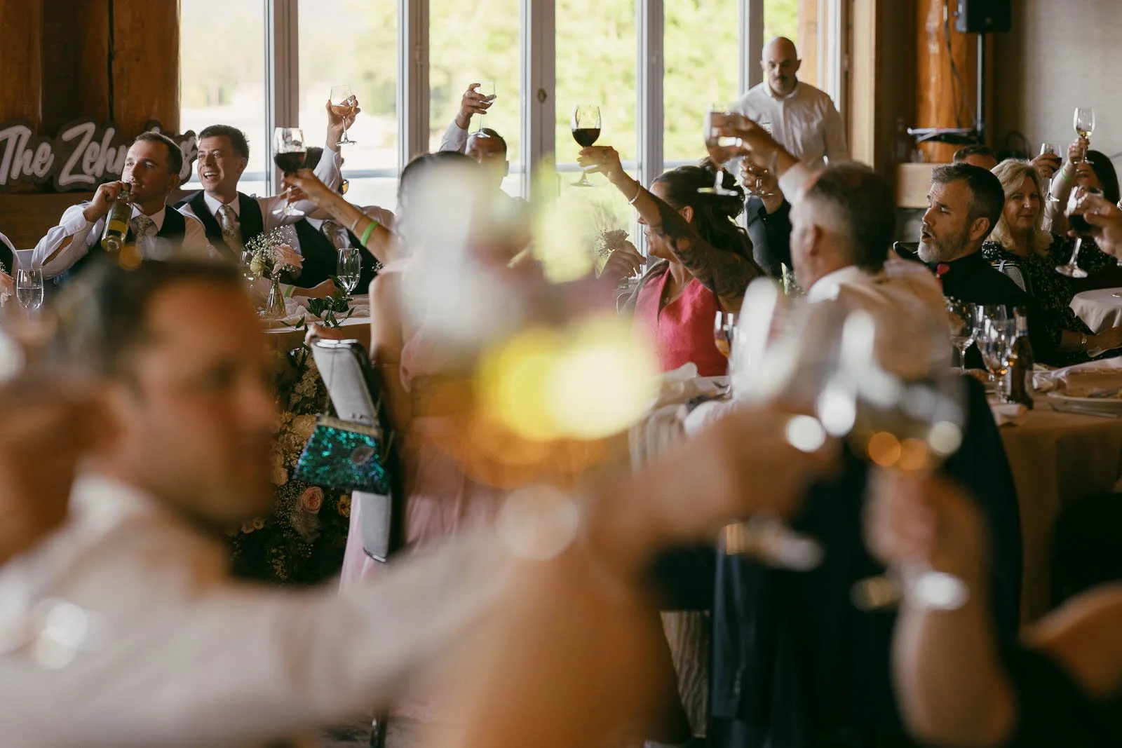  A group of people seated at round tables indoors raise their glasses in a toast, with warm sunlight streaming through large windows in the background. 
