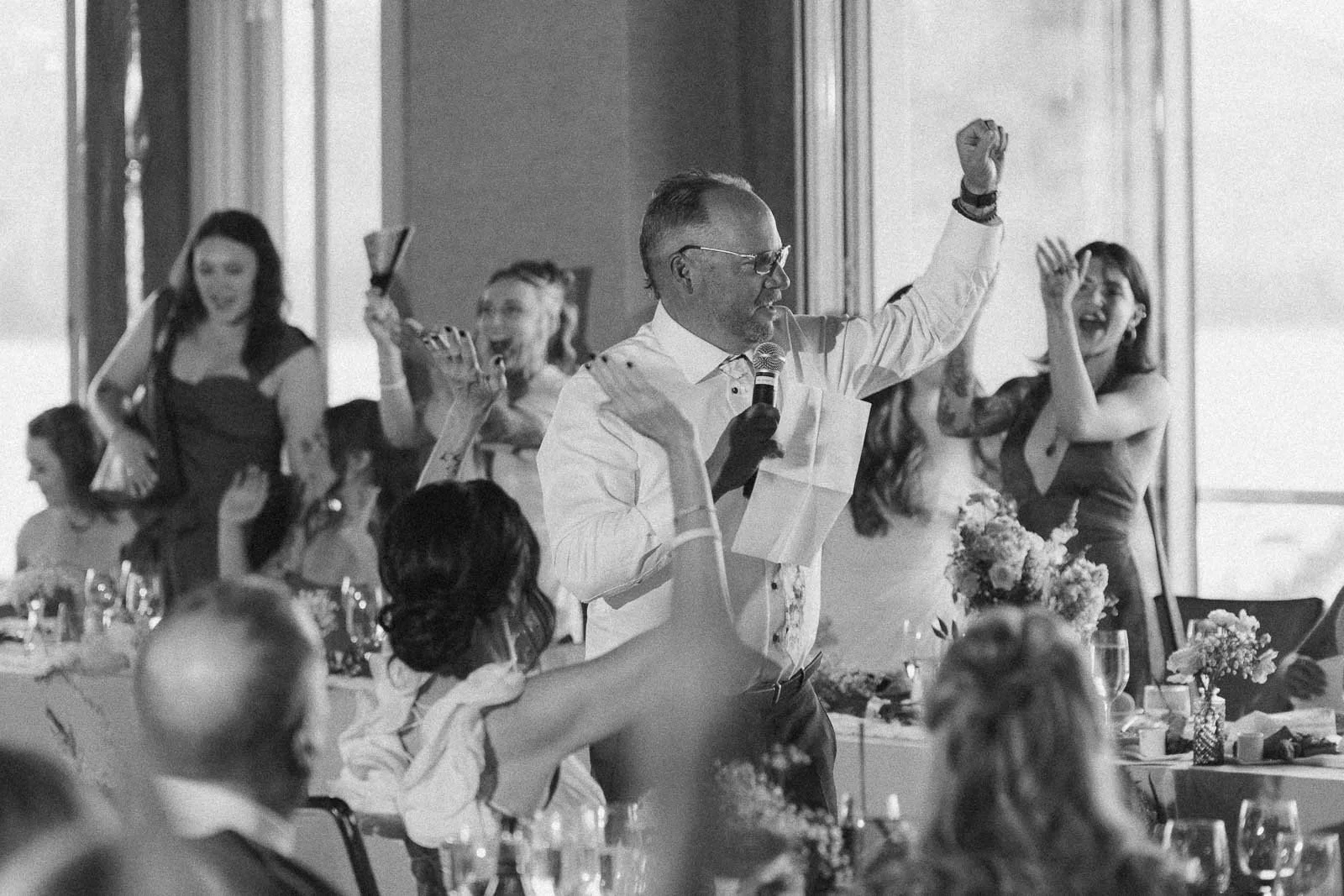  The father of the bride holding a microphone and paper gives a toast at a lively event as guests cheer and raise their arms in celebration around him. 