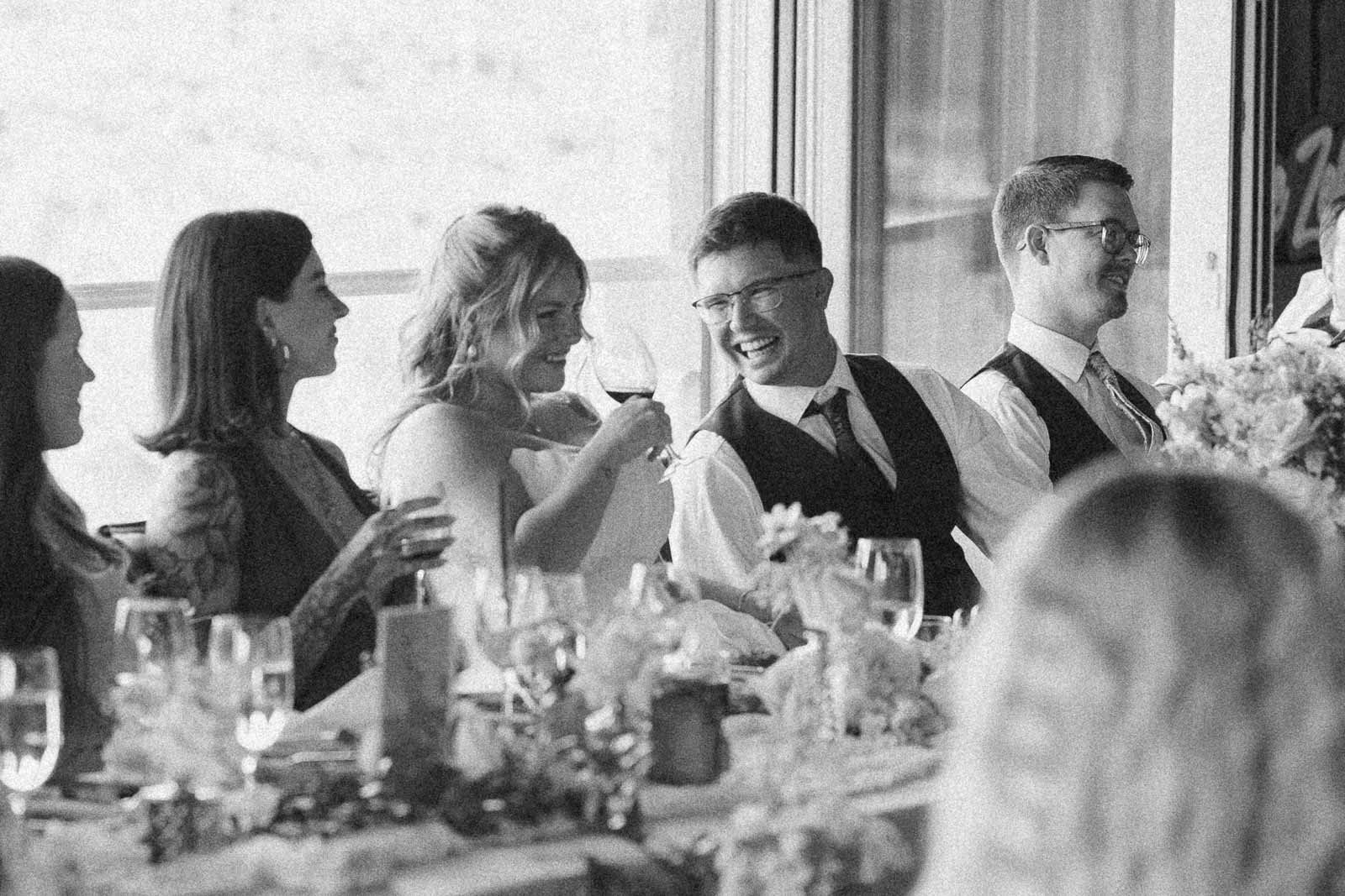  A group of people dressed formally sit at a decorated table, smiling and raising glasses in a celebratory toast. The scene has a joyful, candid atmosphere. 