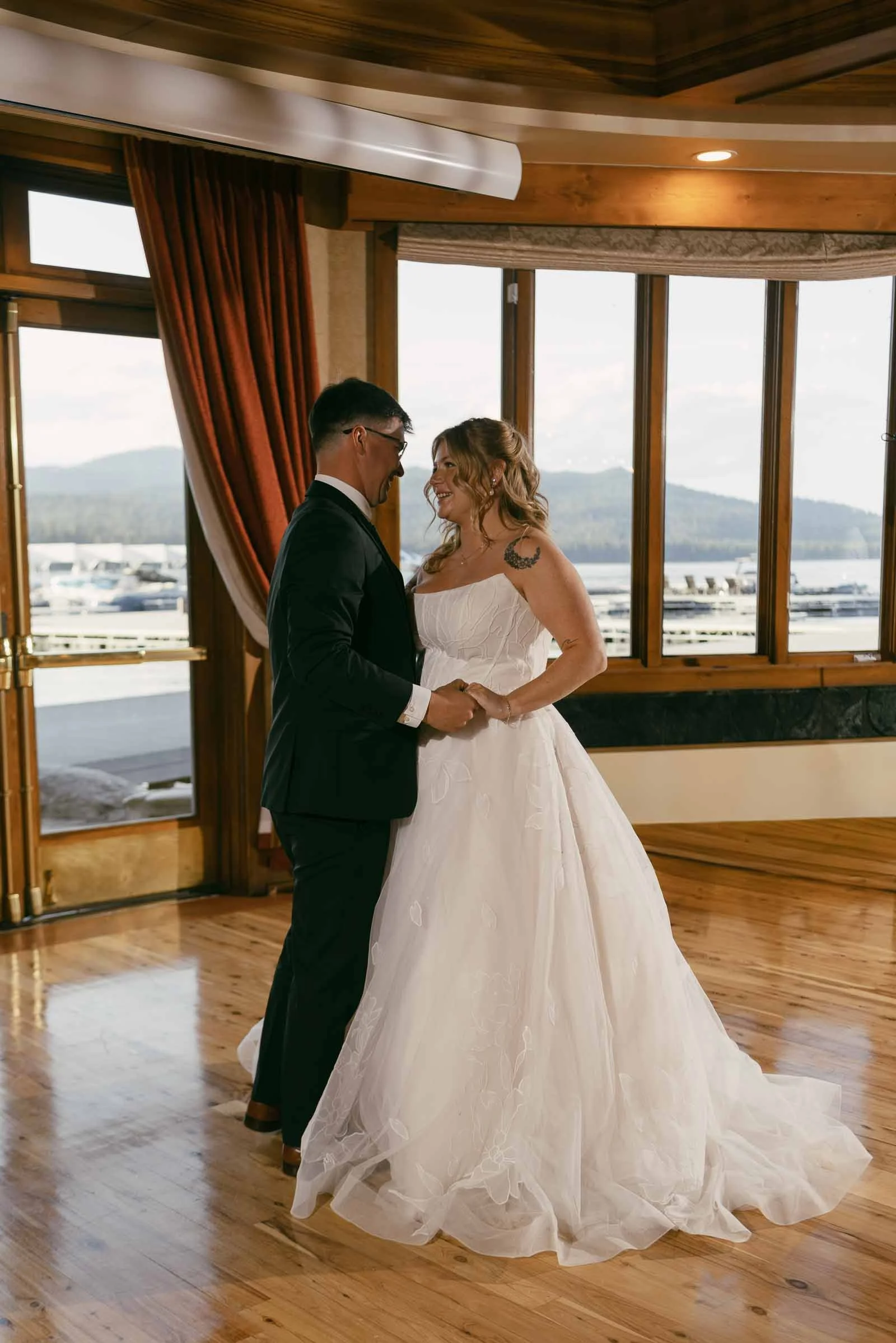  A bride and groom dance together in a sunlit room with wooden floors, large windows, and mountains visible outside. 