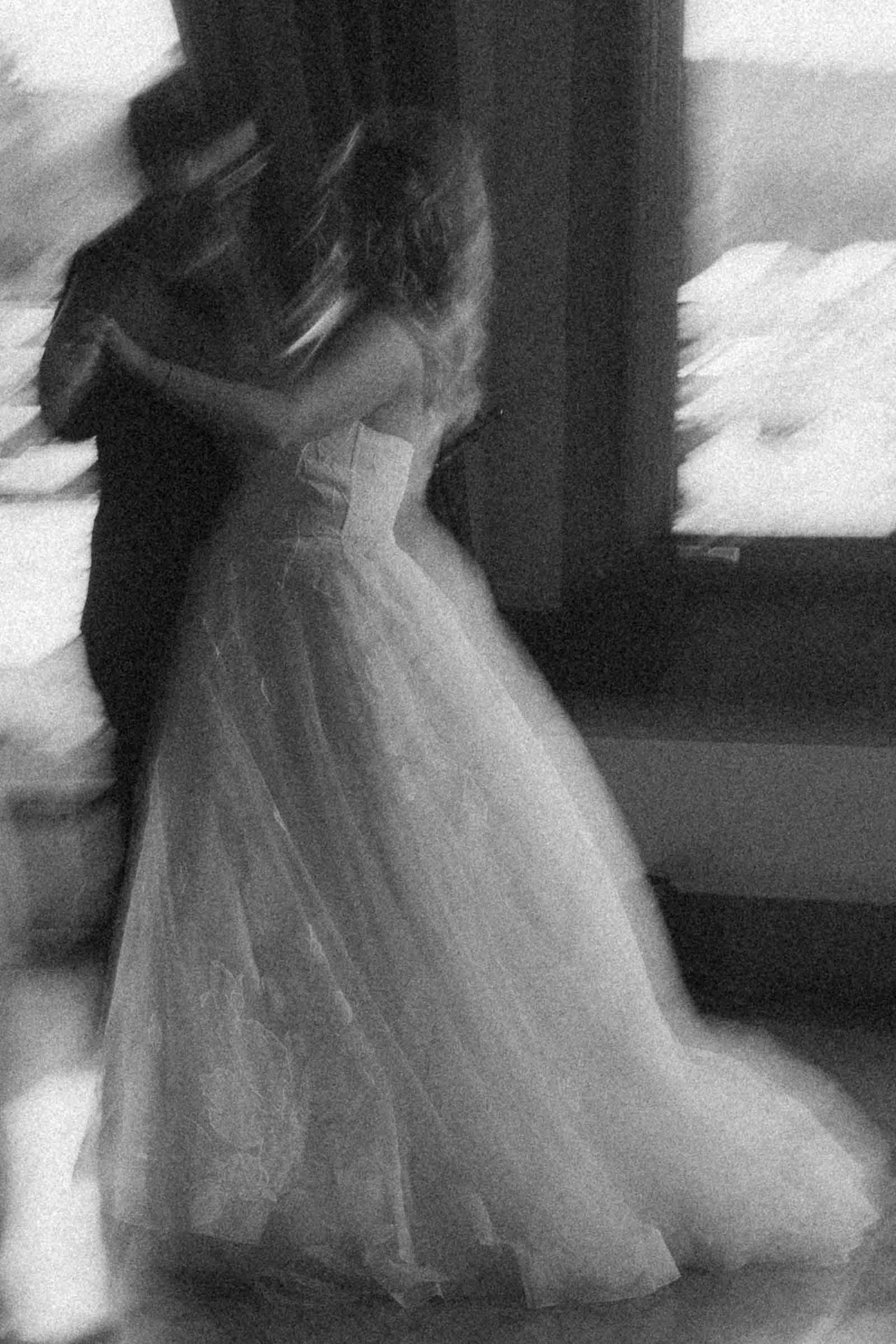 A black and white, blurry image of a couple dancing. The woman wears a long, flowing dress and the man is in dark attire. They appear to be inside, near large windows, creating a dreamy, romantic atmosphere. 