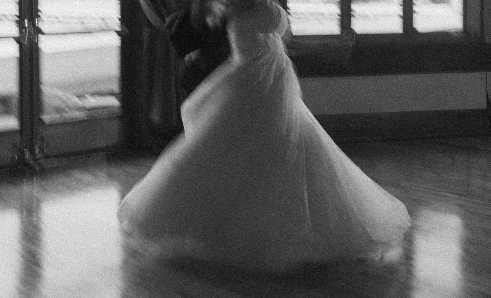  A blurred black and white photo of a couple dancing, with the woman in a flowing wedding dress and the man in a suit, spinning together on a polished floor near large windows. 