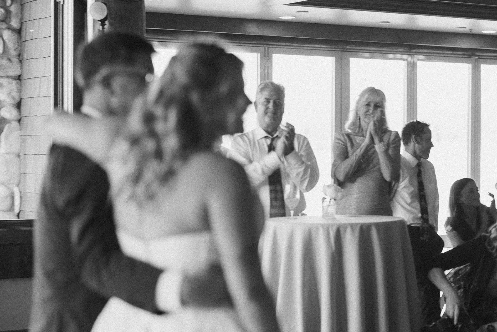  A blurred couple dances in the foreground while four people stand by a round table, clapping and smiling, inside a sunlit room with large windows. 