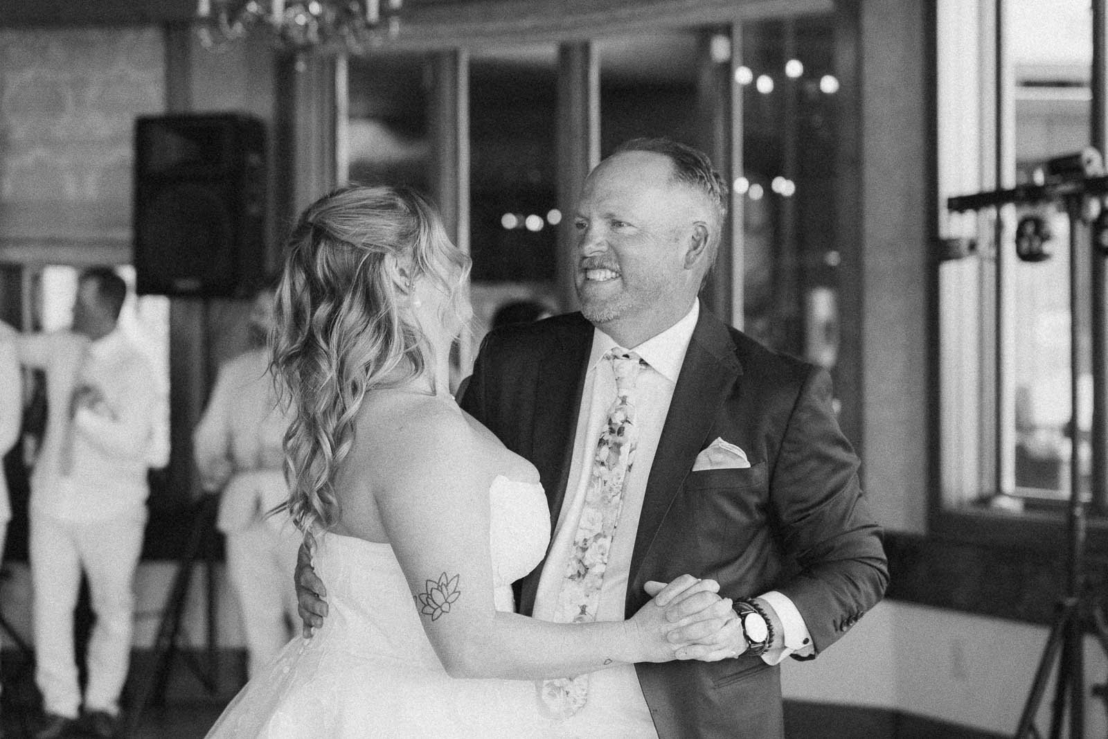  A bride and her father smile at each other while dancing together indoors, surrounded by wedding guests. 