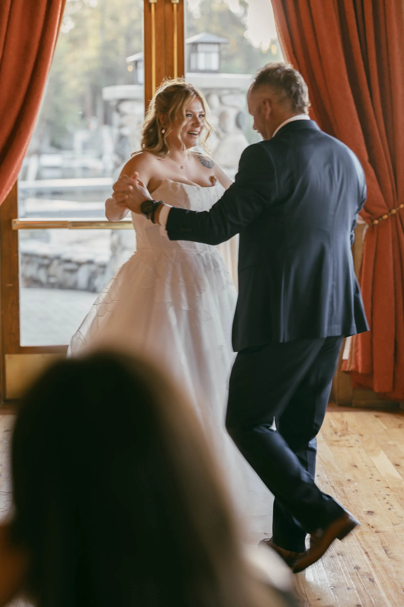  A bride in a white dress and her dad dressed in a suit dance together in a warmly lit room with red curtains and large windows, smiling and enjoying the moment. 