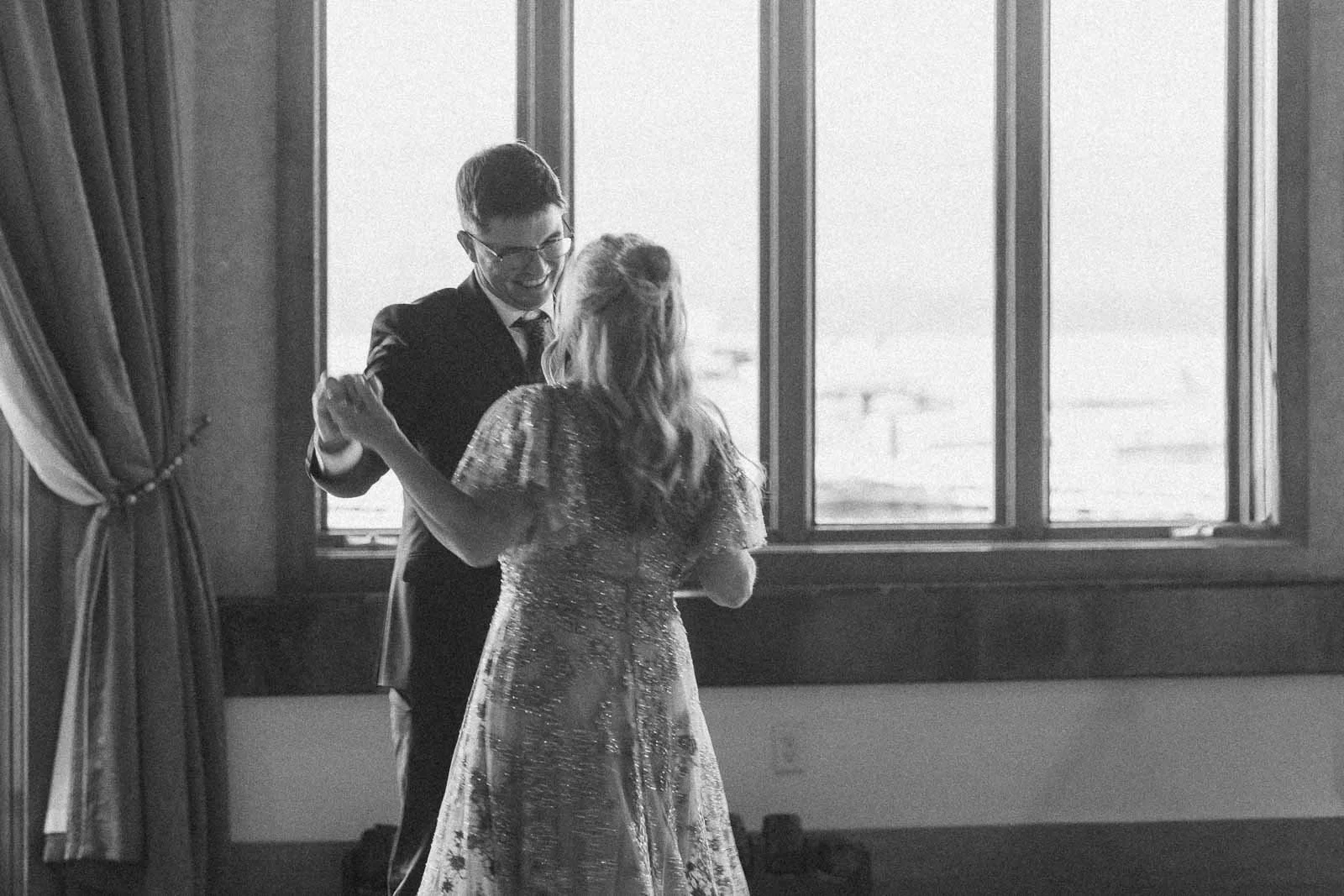  A groom and his mom dance together in front of large windows, with soft natural light illuminating the room. The scene has a joyful, intimate atmosphere. 