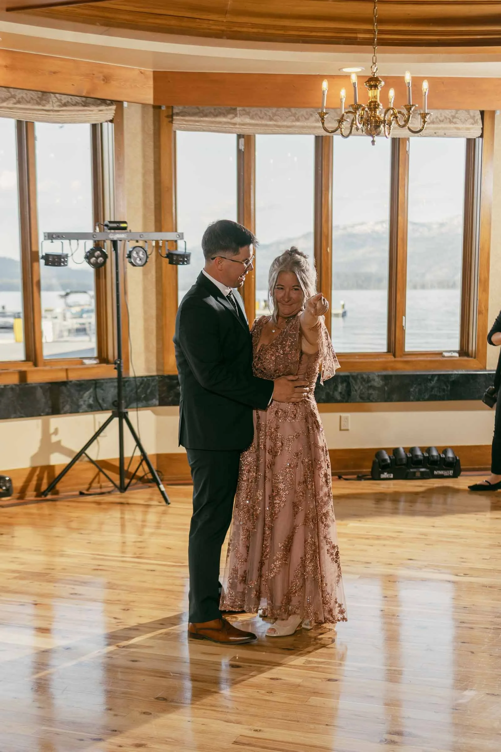  A groom in a suit dances with his mom in a pink, floral dress on a wooden floor in a room with large windows, mountain views, and a chandelier above. 